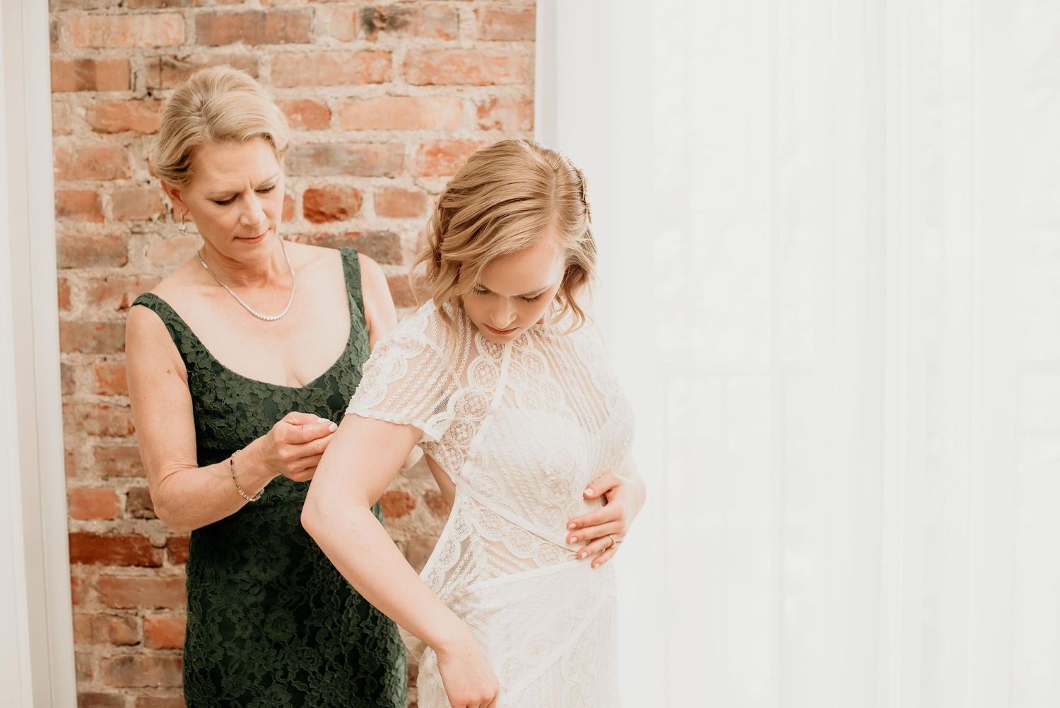 Mother helping bride button up lace wedding dress in front of brick wall and white curtain.
