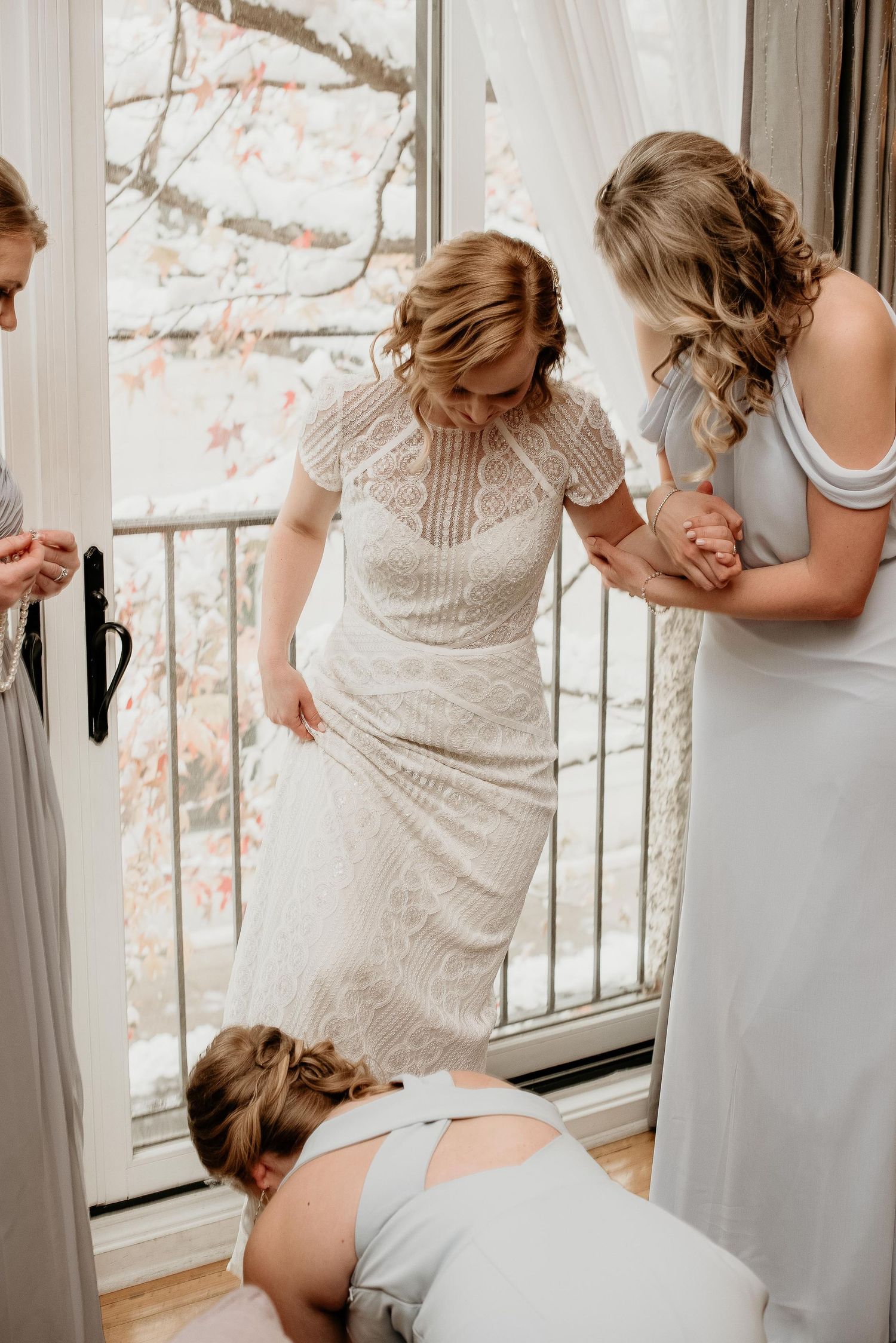 Bridesmaids helping bride into white lace wedding dress near a bright window with autumn foliage outside.