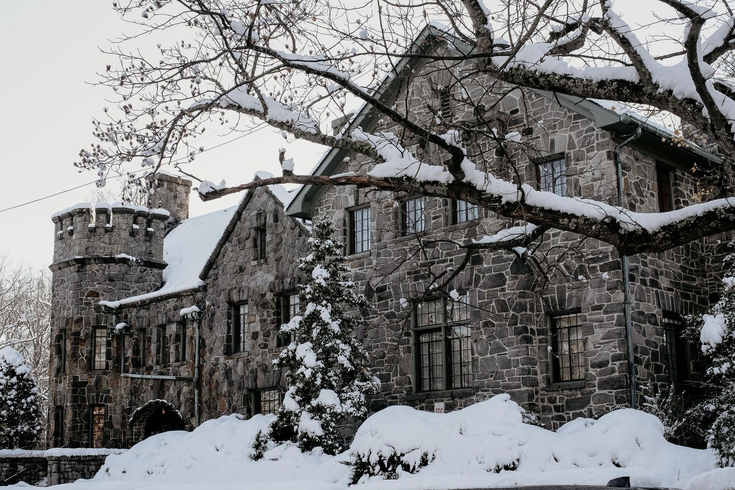 Snow-covered stone castle with tower and evergreen trees in winter landscape.