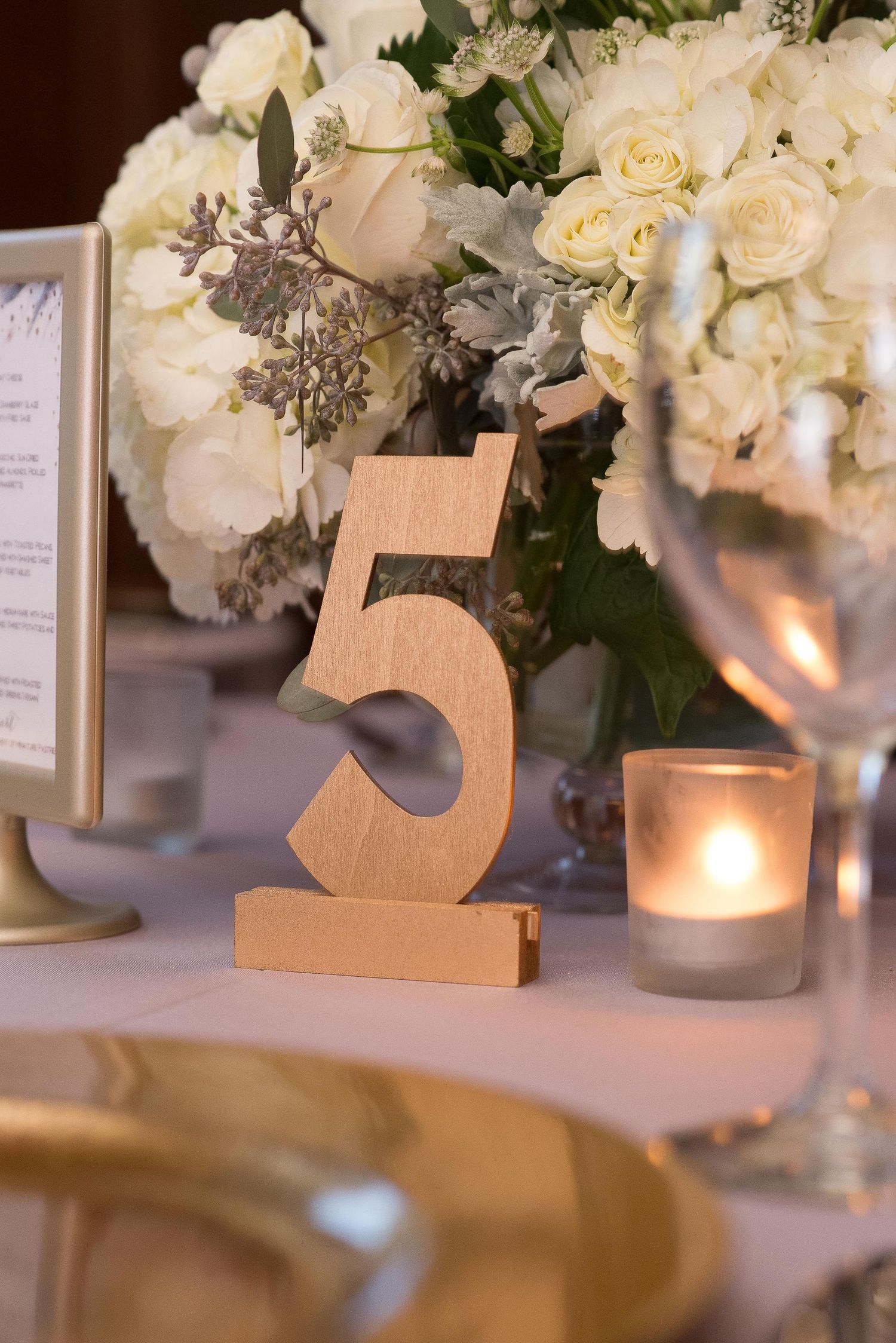 Gold wooden number 5 table sign at elegant wedding reception with white roses centerpiece and candlelight.