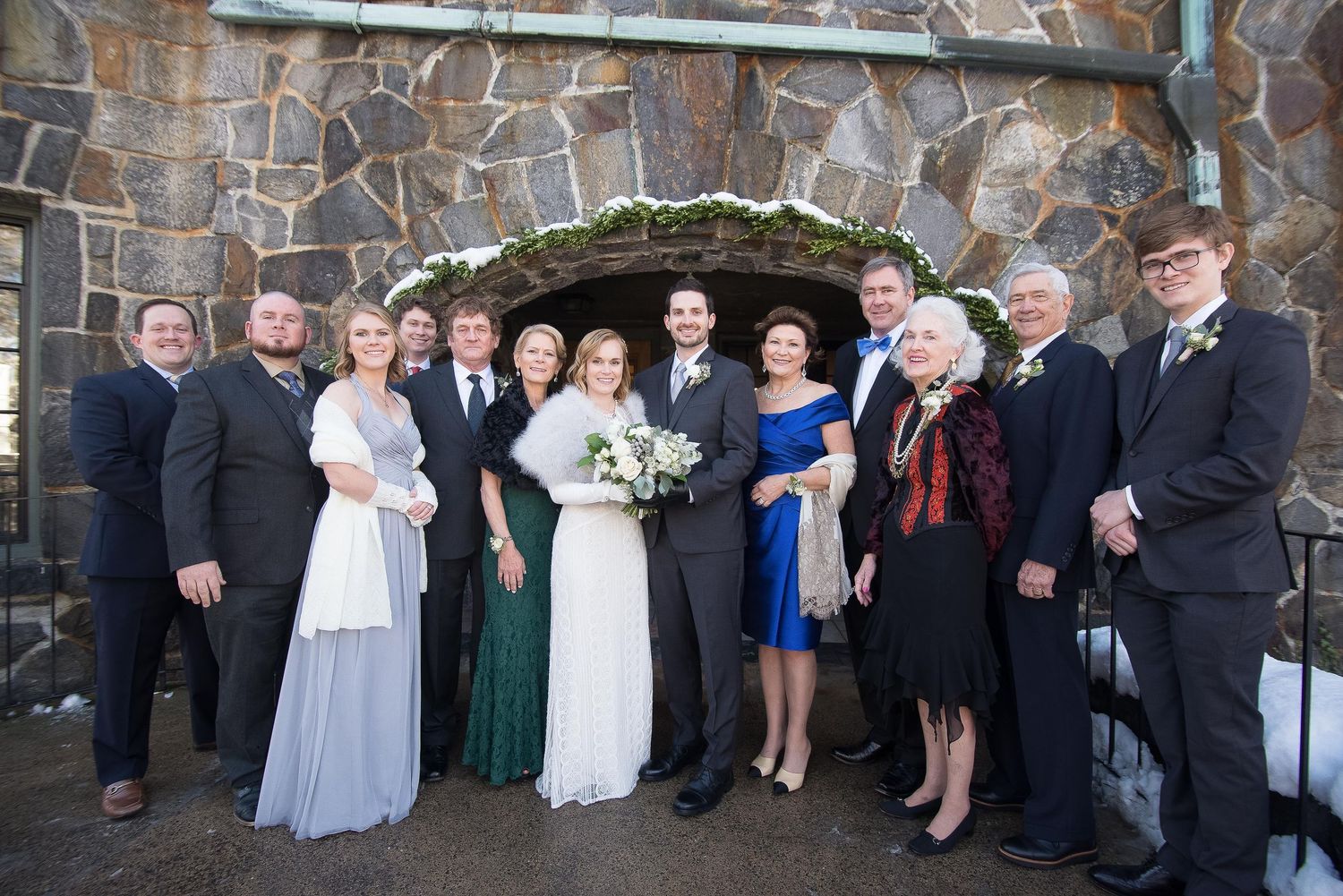 Wedding party poses in front of stone arch, bride in white gown with bouquet, guests in formal attire outdoors.