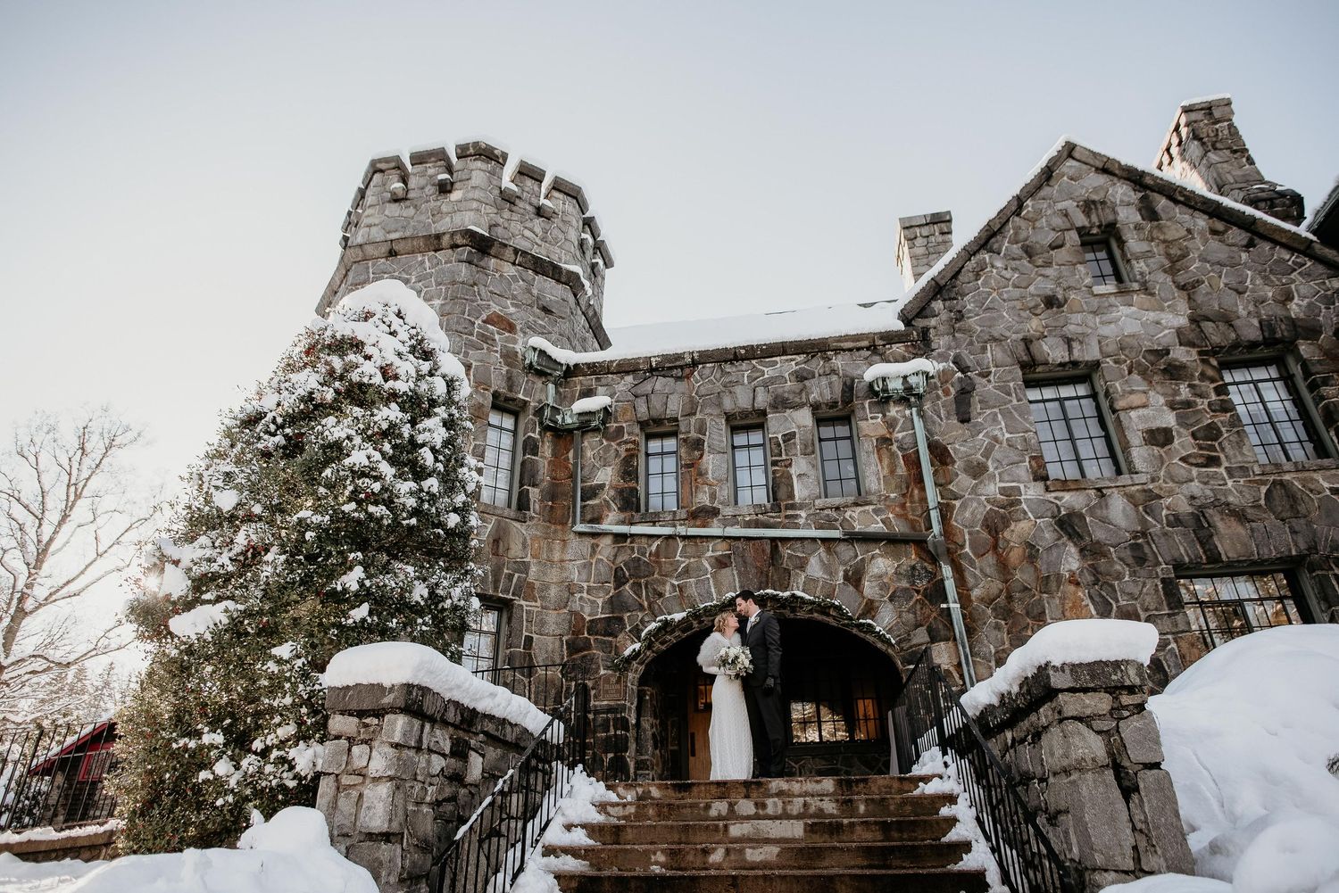 Bride and groom embracing at snow-covered stone castle entrance in winter wedding portrait.