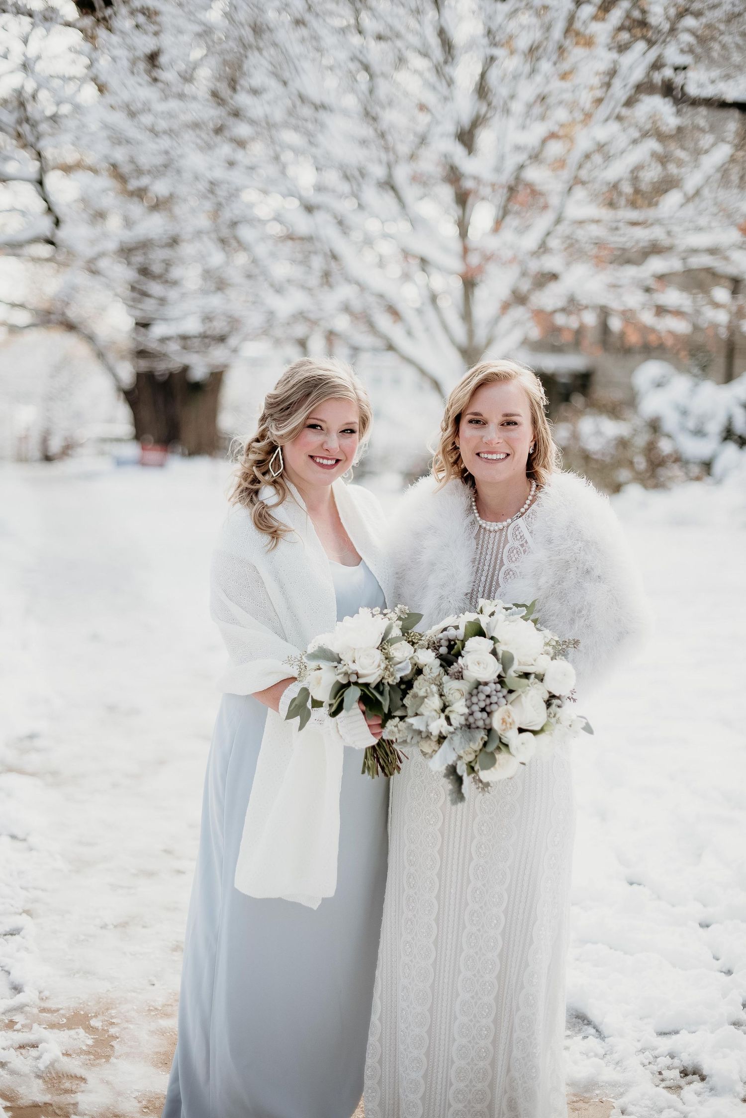 Two brides in elegant winter wedding attire holding white bouquets, posing outdoors in a snowy landscape.