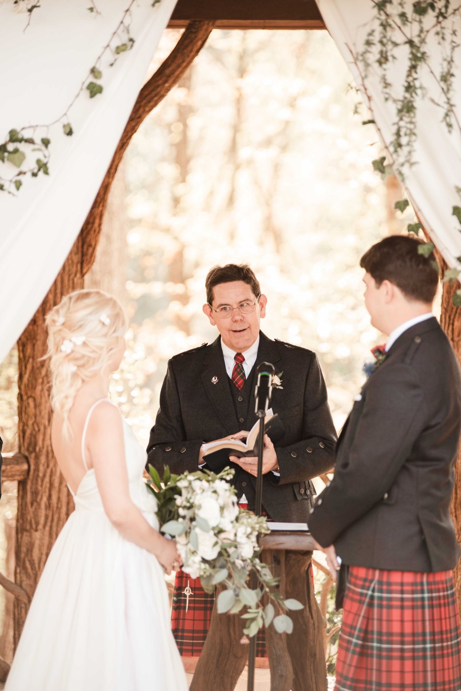 Outdoor wedding ceremony with bride holding bouquet, groom in tartan kilt, and officiant at rustic wooden altar.
