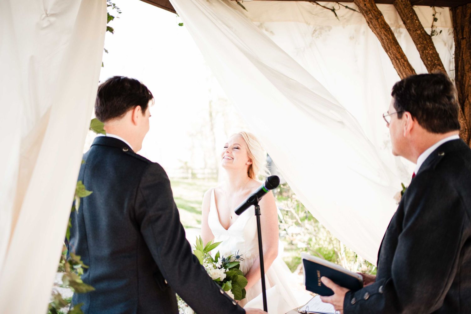 Bride smiling at groom during outdoor wedding ceremony with officiant and microphone under white draped canopy.
