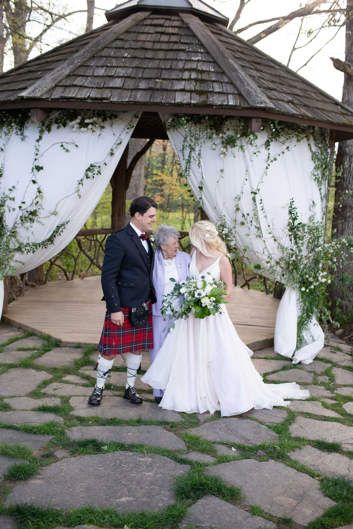 Bride and groom in Scottish kilt stand with elderly guest under floral gazebo on stone path at outdoor wedding.