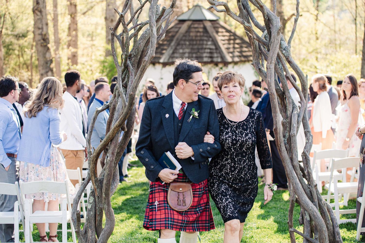 Man in Scottish kilt and woman in lace dress walk arm-in-arm at outdoor wedding ceremony under rustic arch.