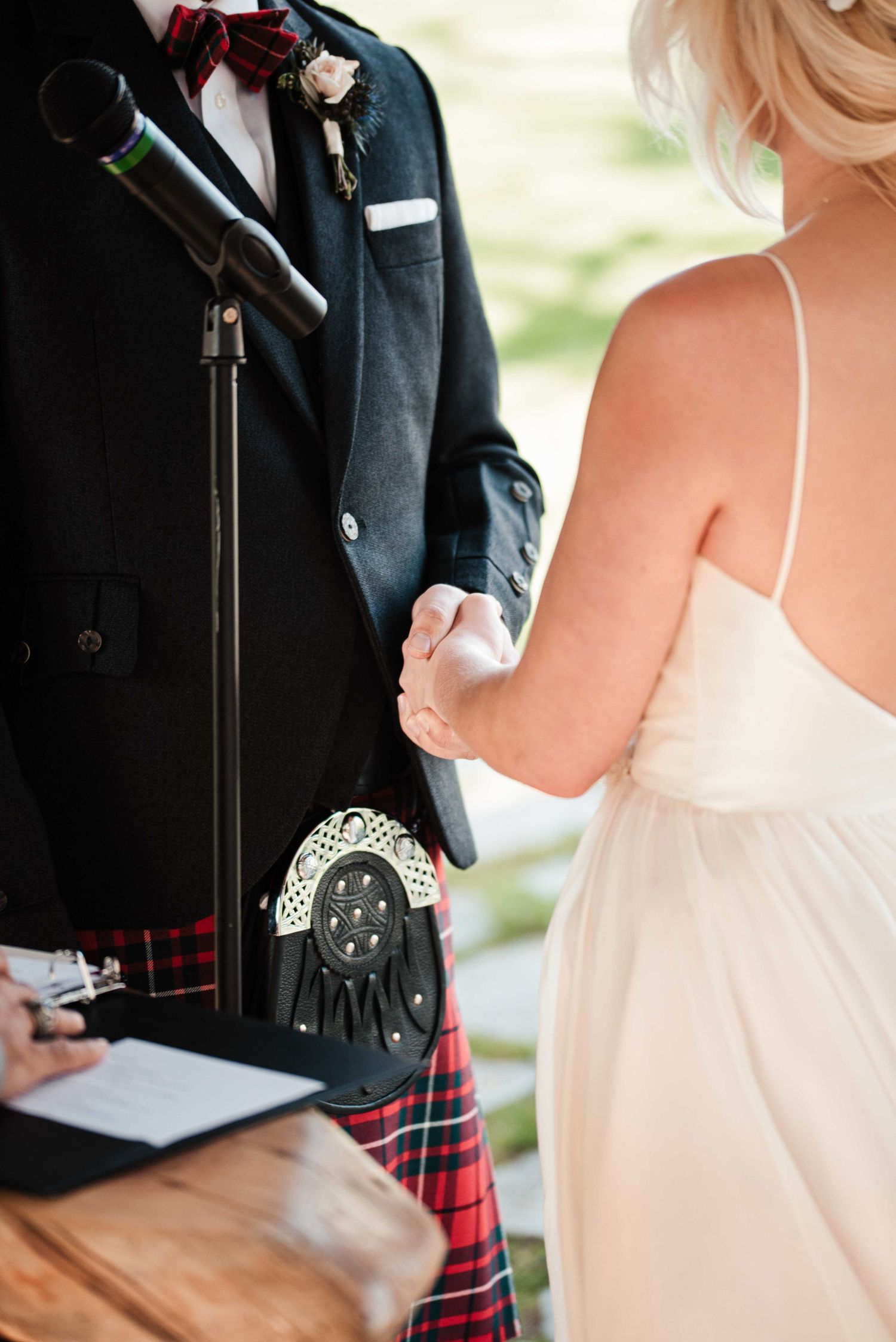 Groom in Scottish kilt and bow tie holds bride's hand during outdoor wedding ceremony.