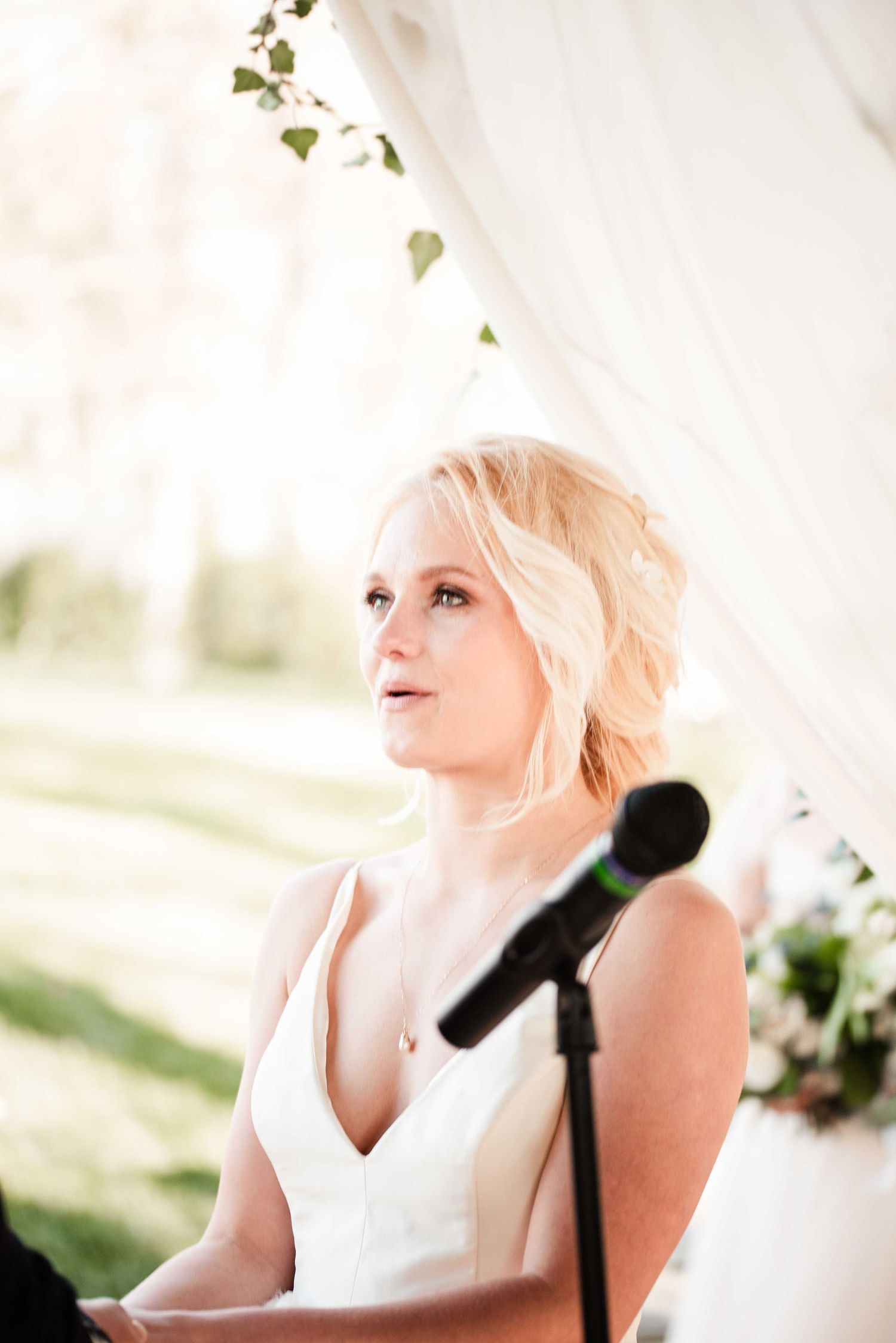Blonde bride in white gown gazes upward near microphone stand during outdoor wedding ceremony.