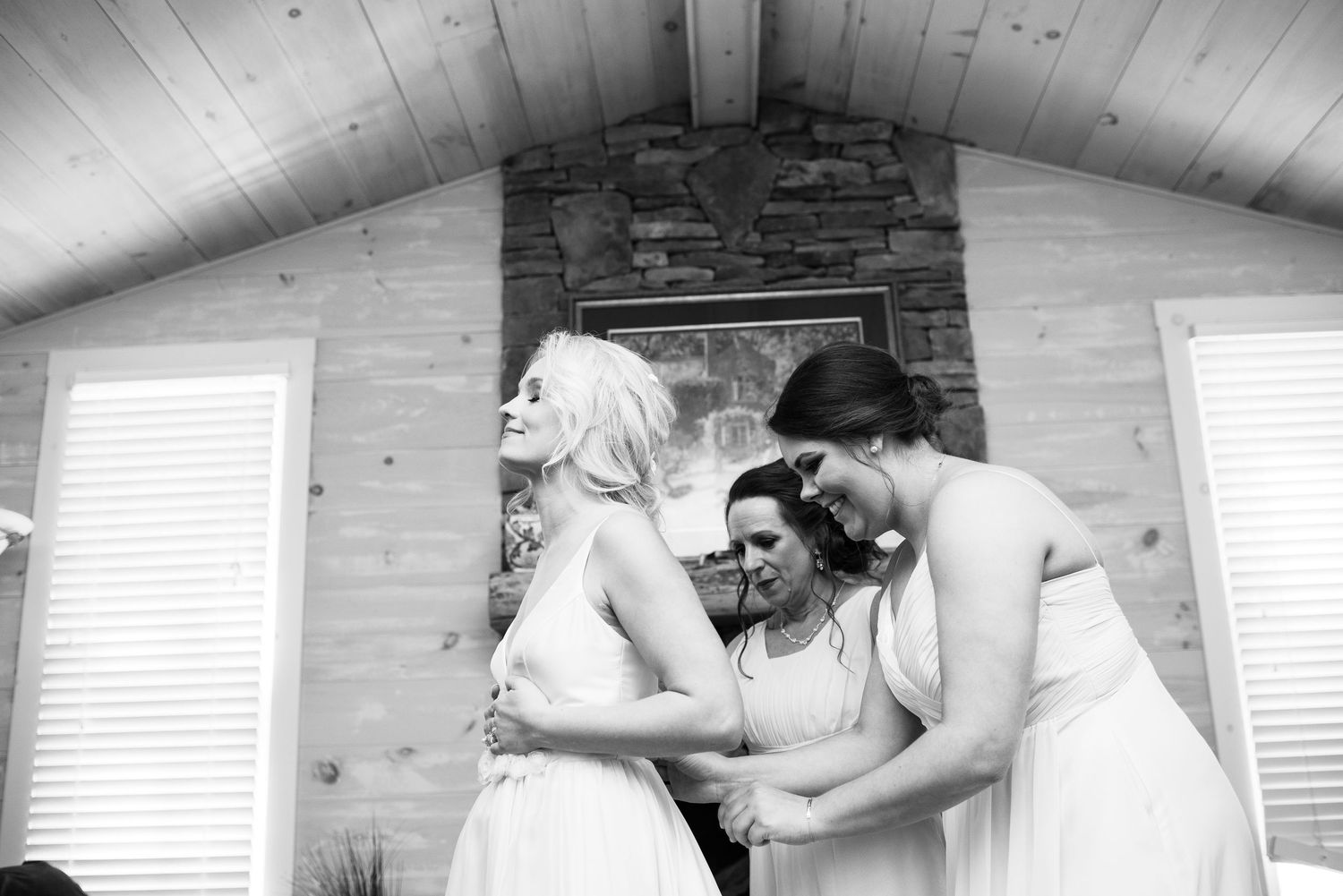 Black and white photo of bridesmaids helping the bride with her dress inside a wood-paneled chapel.