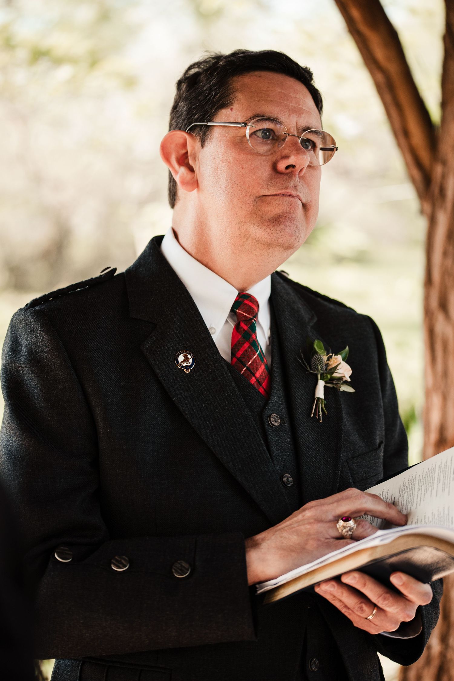 Officiant in dark suit with tartan tie holds book during outdoor wedding ceremony.