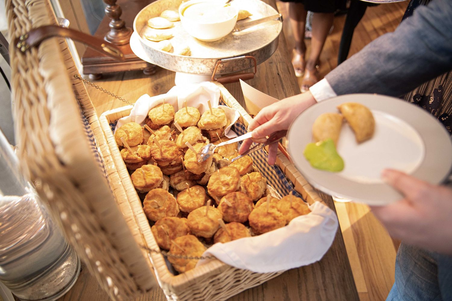 Person serving freshly baked golden muffins from a wicker basket lined with white cloth at a buffet spread.