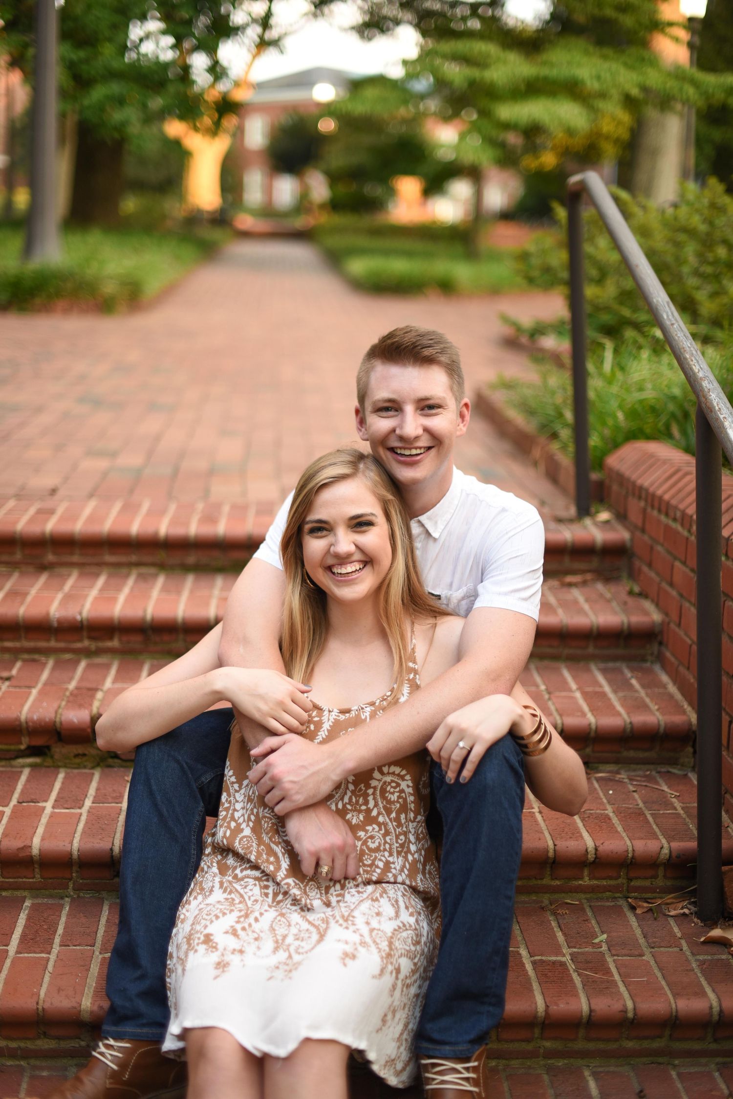 Smiling couple sits on brick steps outdoors, man embracing woman from behind in a garden setting.