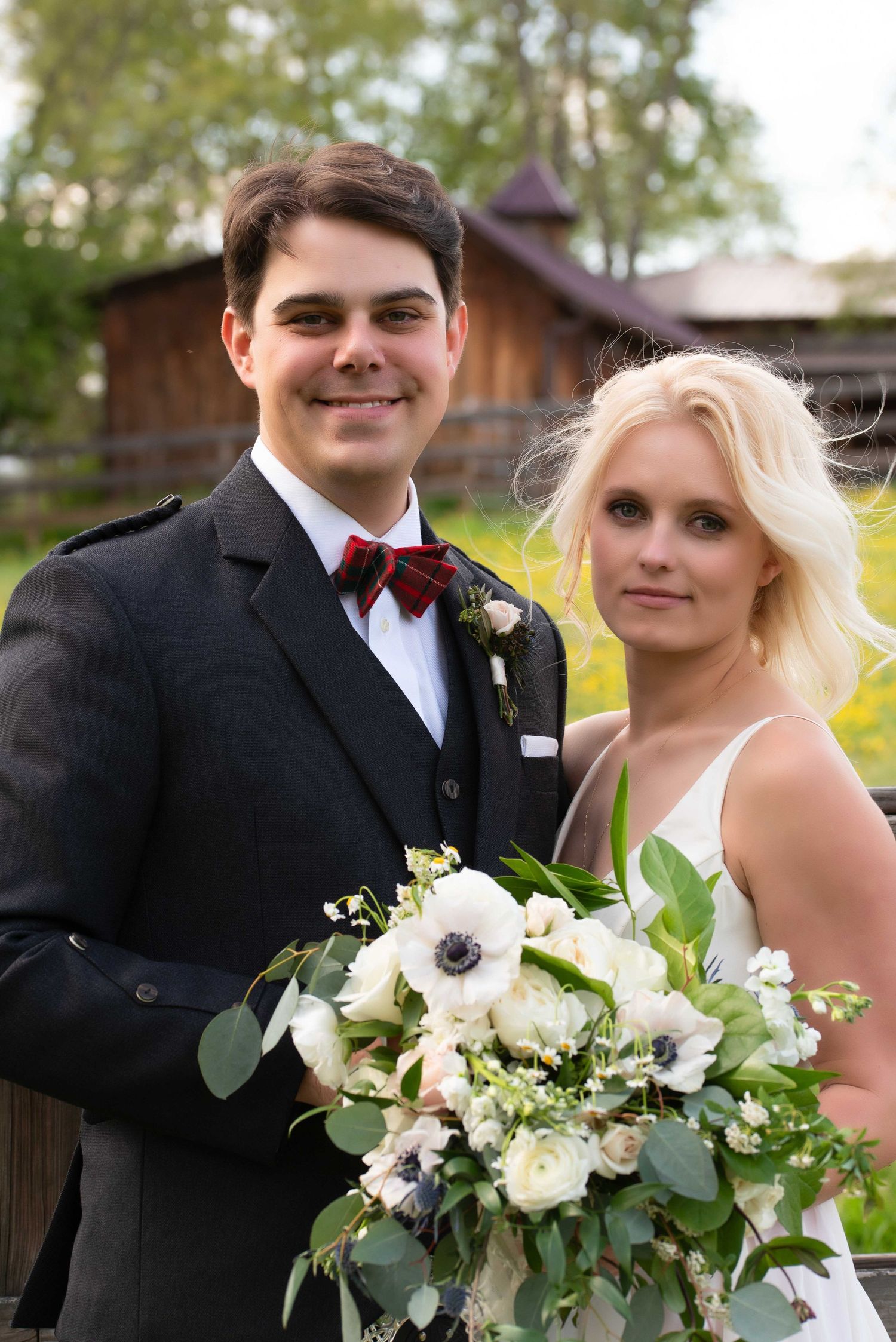 A smiling couple in wedding attire poses outdoors, groom in black suit with red bow tie, bride holding white floral bouquet.