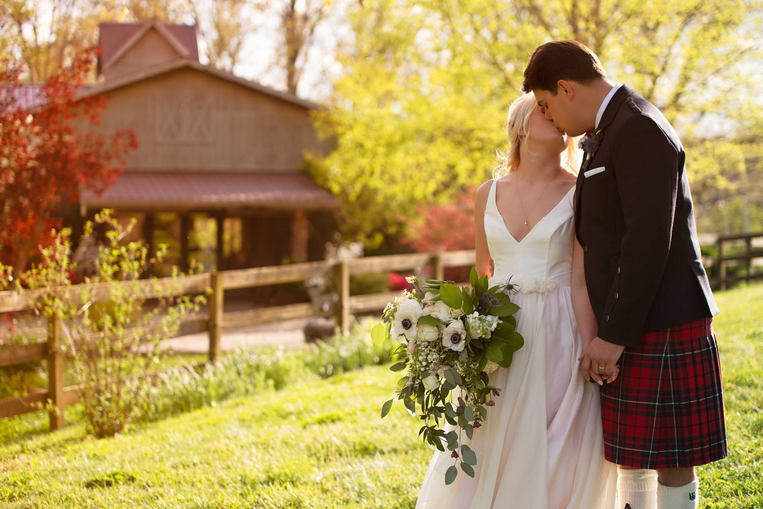 Bride and groom kissing outdoors, groom wearing red tartan kilt, bride holding floral bouquet, rustic barn background.