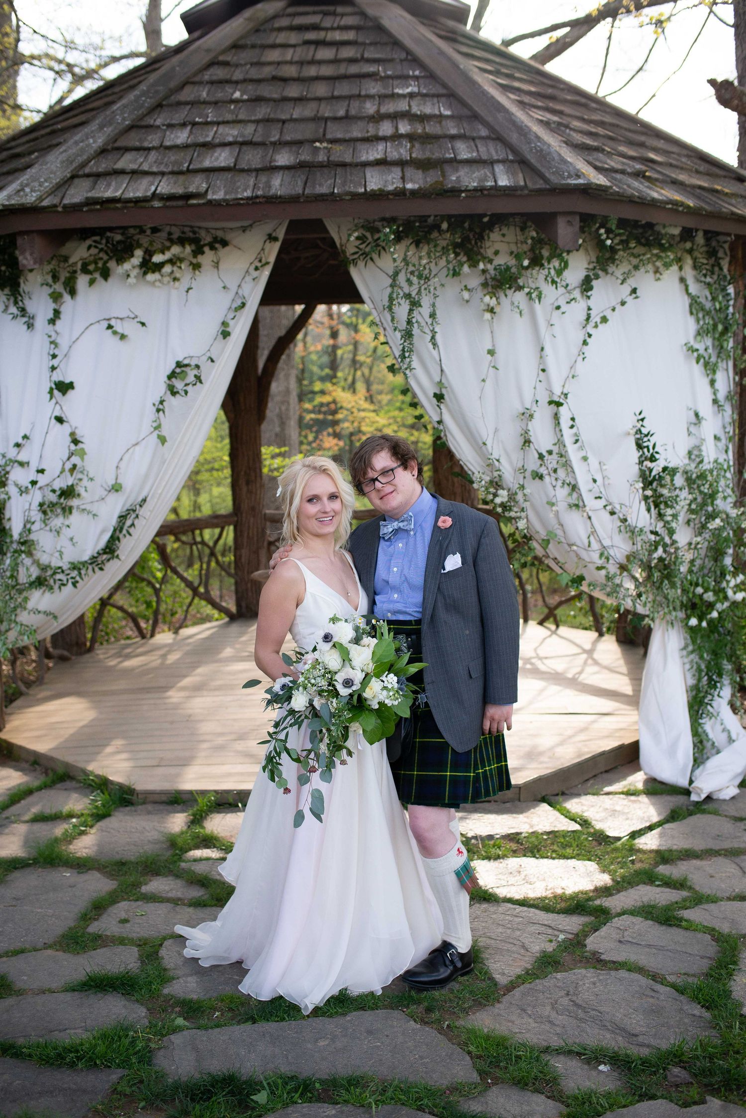 Bride and groom pose before a decorated gazebo with white draping and greenery, bride holding a white bouquet.