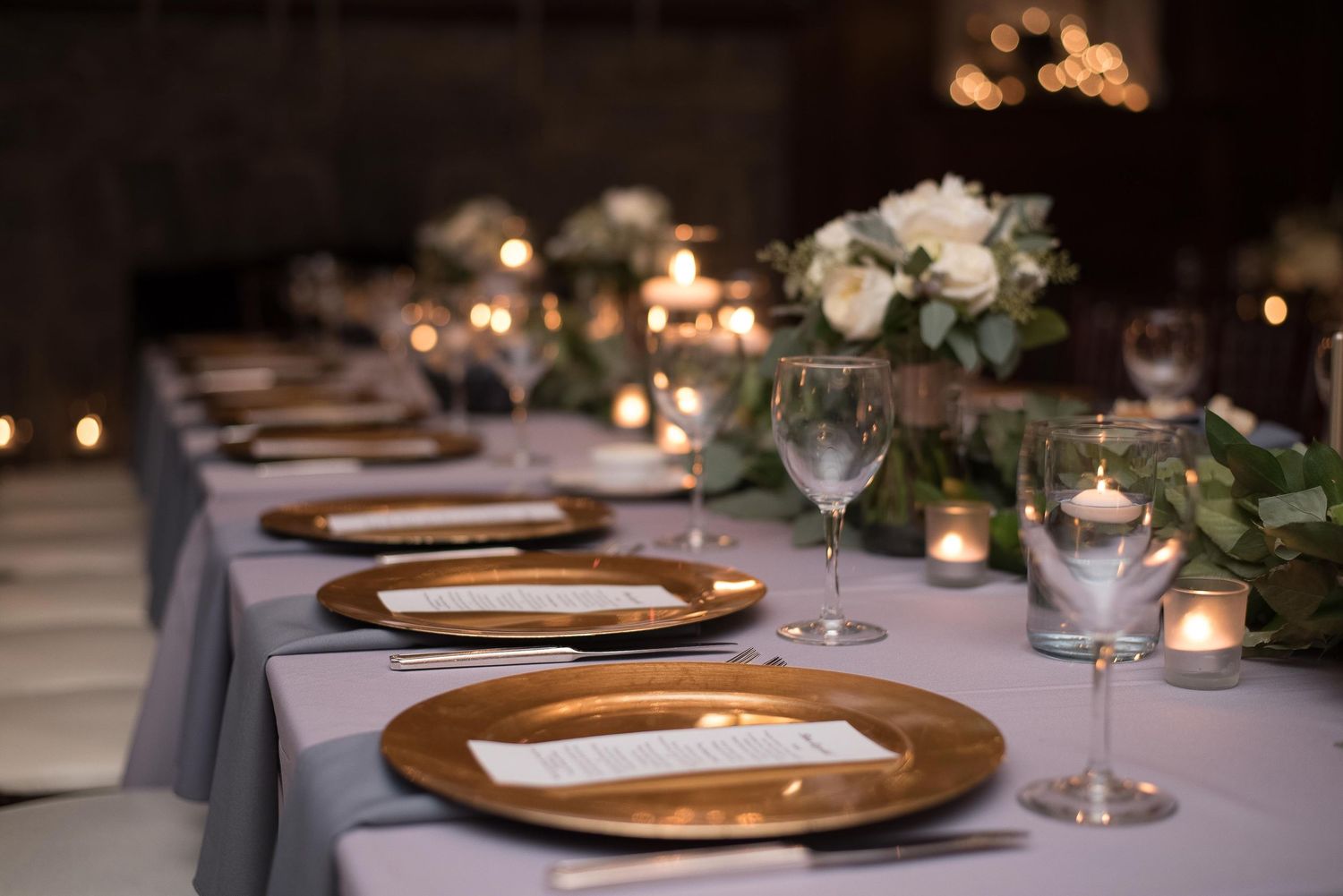 Elegant wedding reception table with gold charger plates, white floral centerpieces, and glowing candles on a gray linen.
