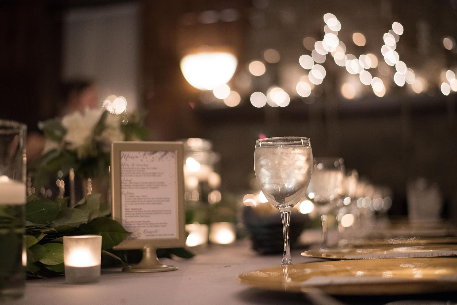Elegant wedding reception table with glowing candles, greenery, wine glasses, and a framed menu card in soft bokeh lighting.