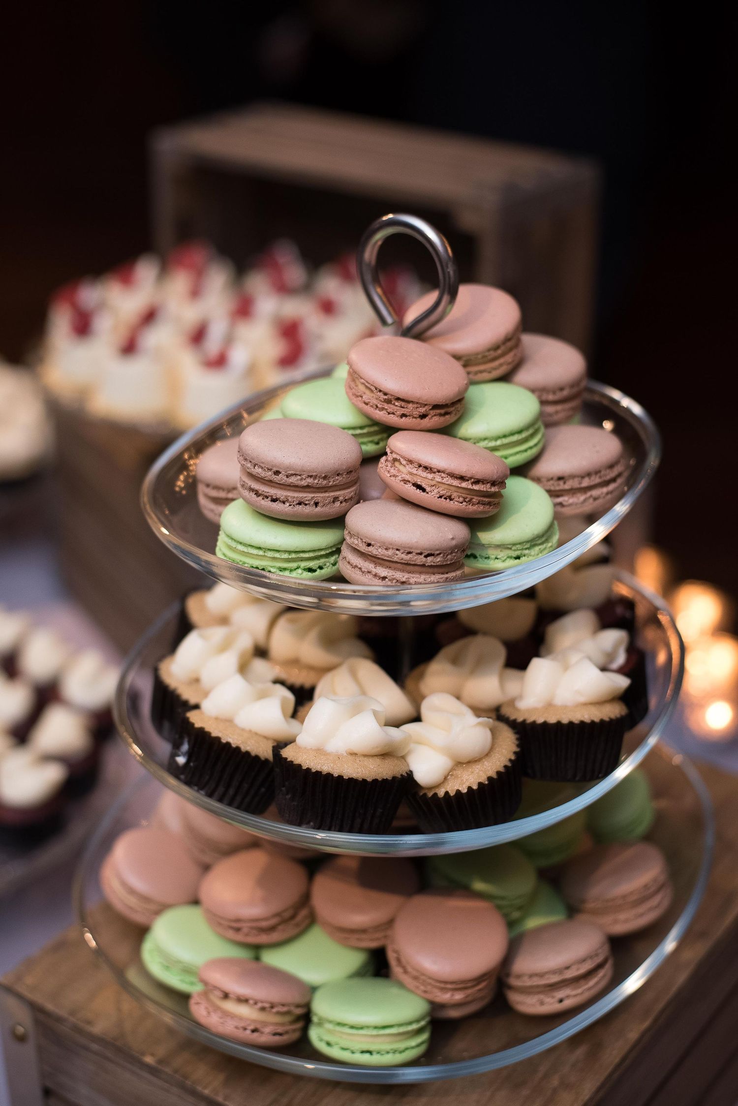 Three-tier glass stand displaying pink and green French macarons and chocolate cupcakes at an elegant dessert table.