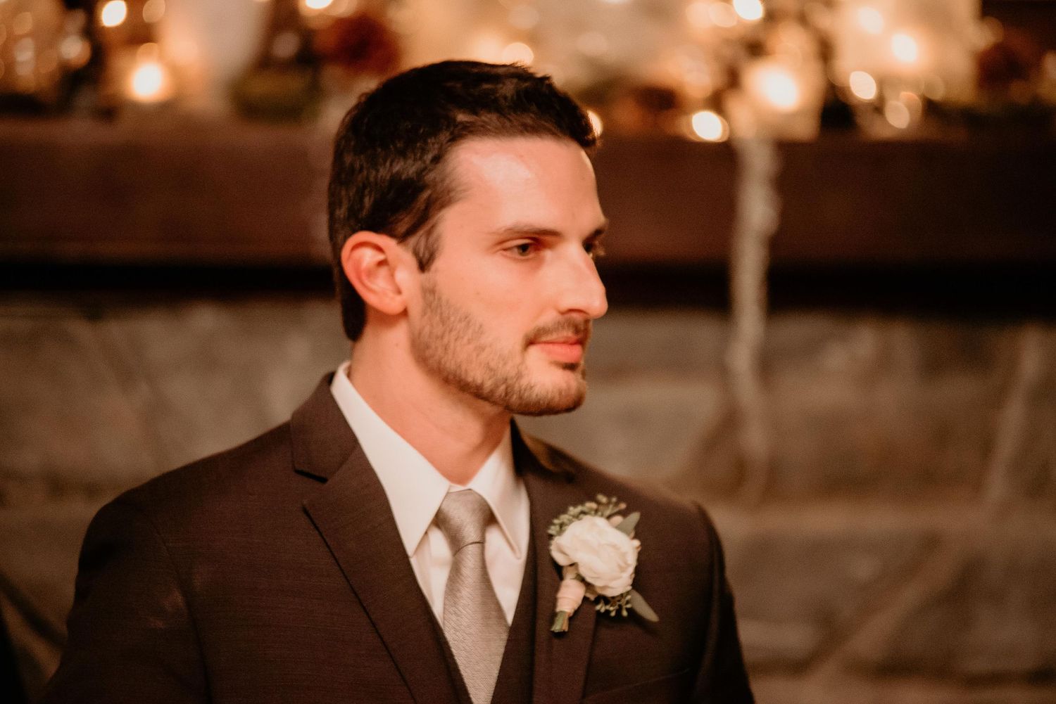 Groom in brown suit with boutonniere standing at wedding altar with warm bokeh lights in background.