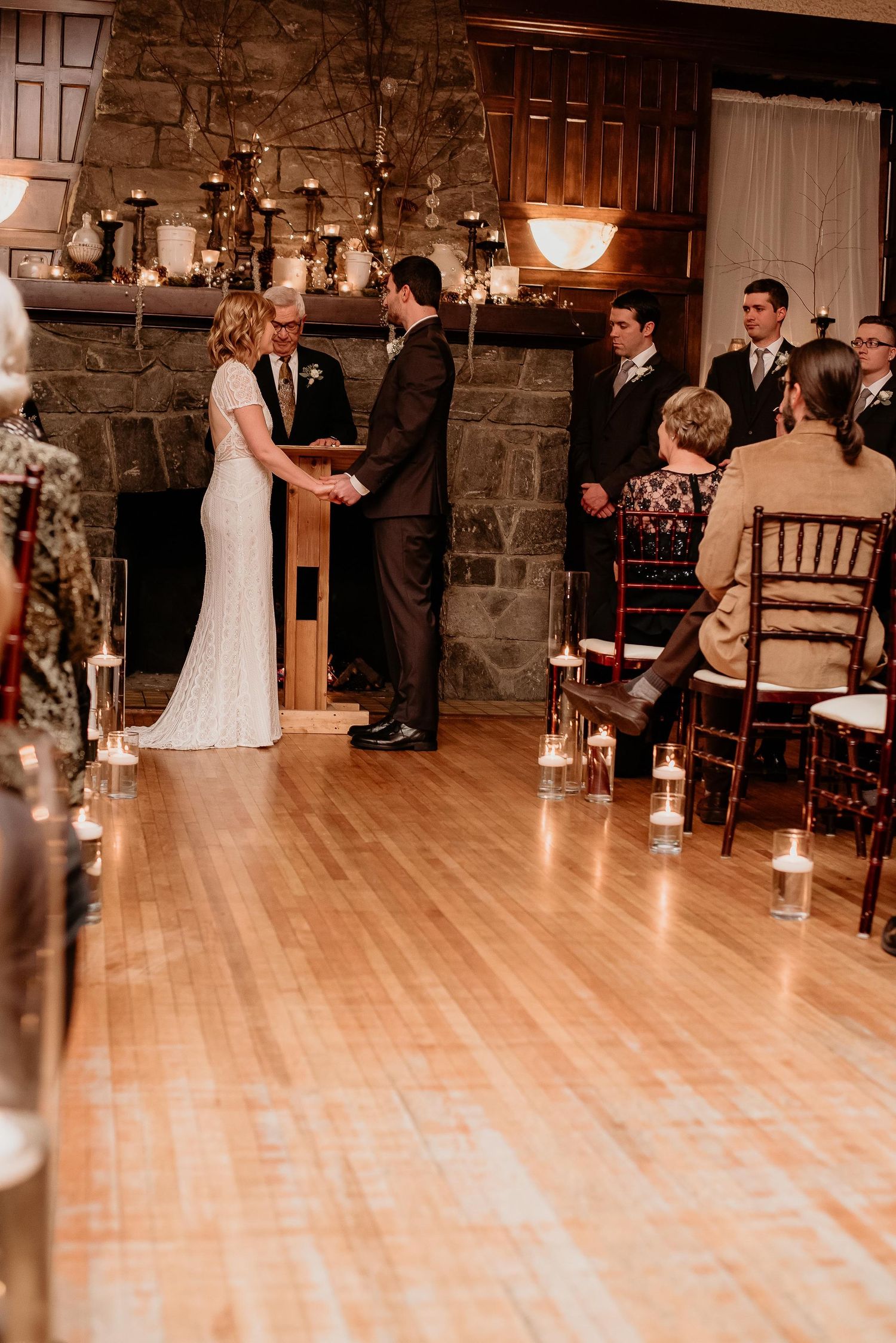 Couple holds hands during indoor wedding ceremony by candlelit stone fireplace with seated guests watching nearby.