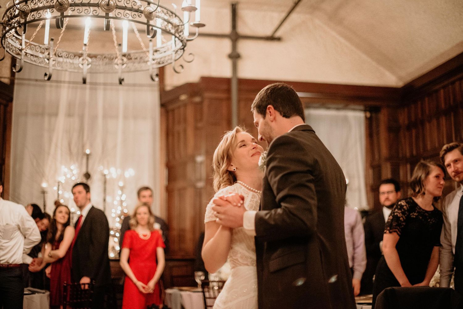 Bride and groom share their first dance in an elegant wood-paneled ballroom with chandelier lighting and guests watching.