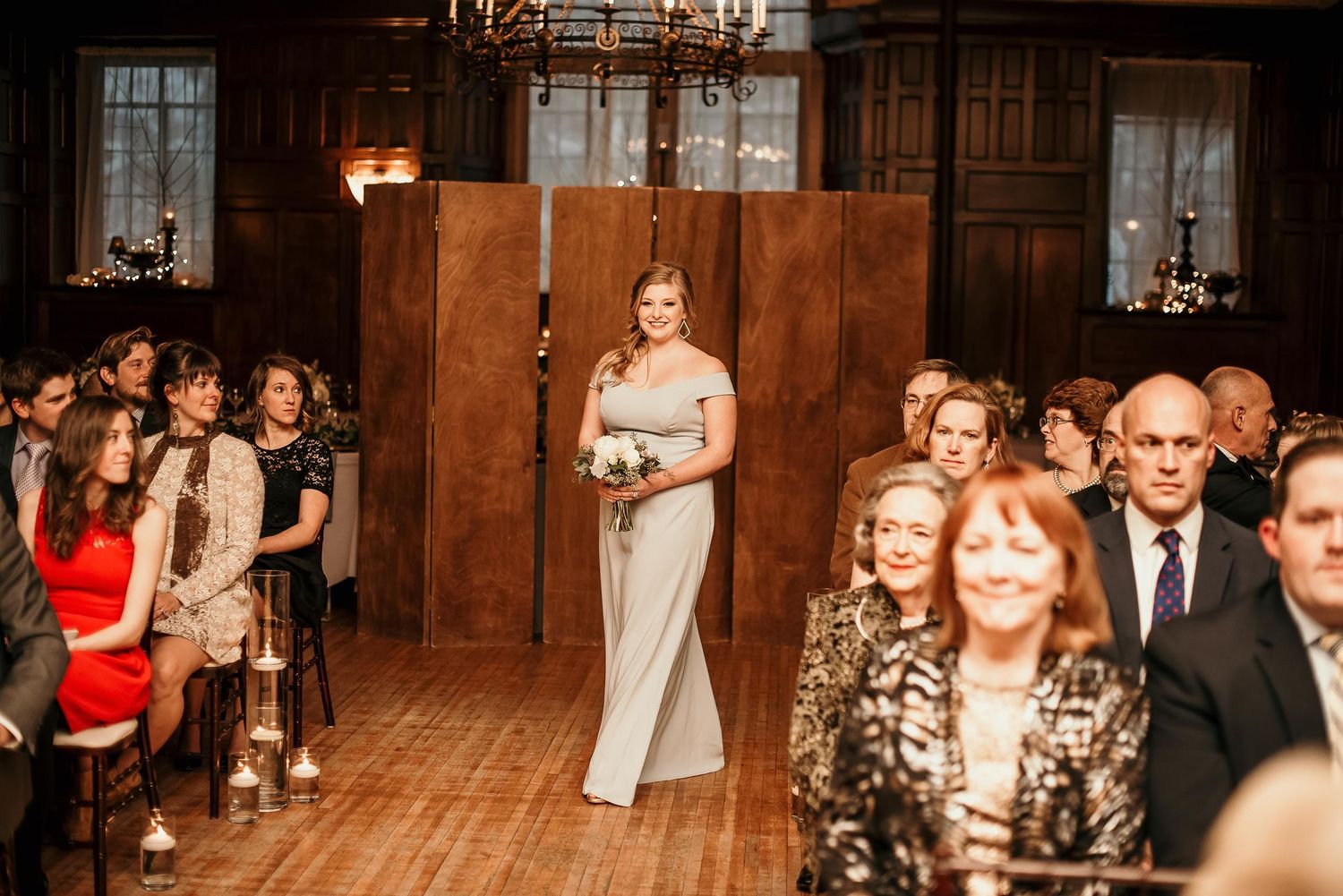 Elegant bridesmaid in silver off-shoulder gown walks down aisle at warmly lit indoor wedding ceremony.