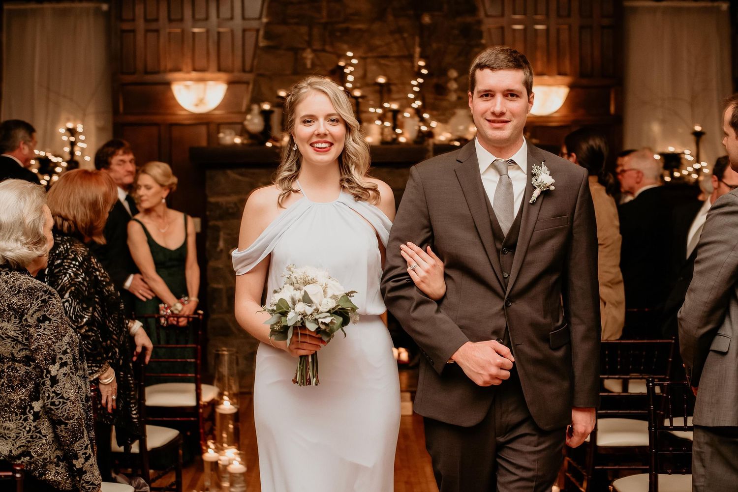 Bride in white gown holding bouquet walks down aisle with groomsman in gray suit at elegant indoor wedding ceremony.