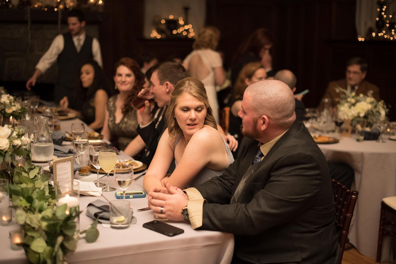 Guests seated at elegantly decorated wedding reception tables, enjoying conversation in a warmly lit venue.