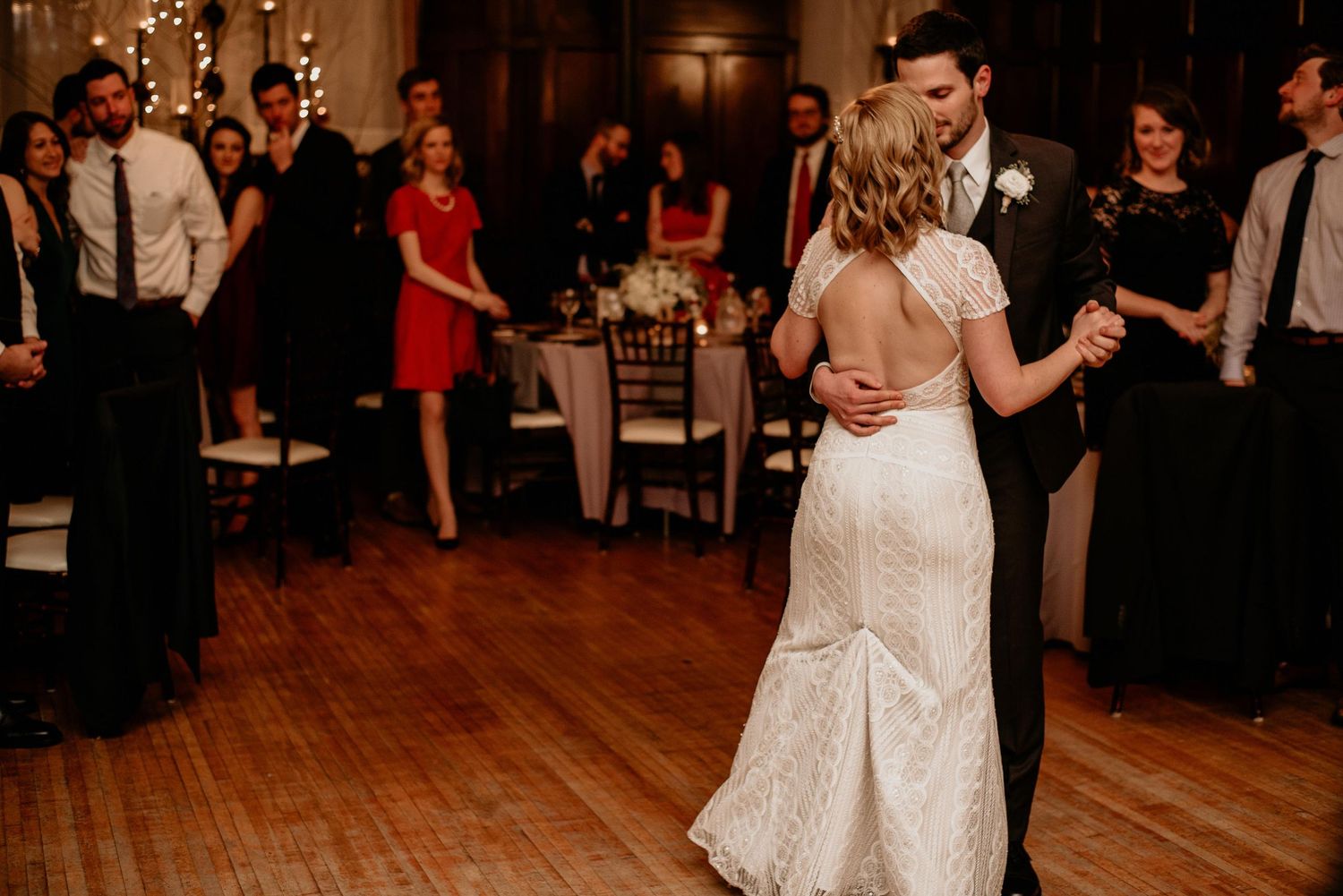 Bride and groom share their first dance at elegant wedding reception with guests watching in warm candlelit ballroom.