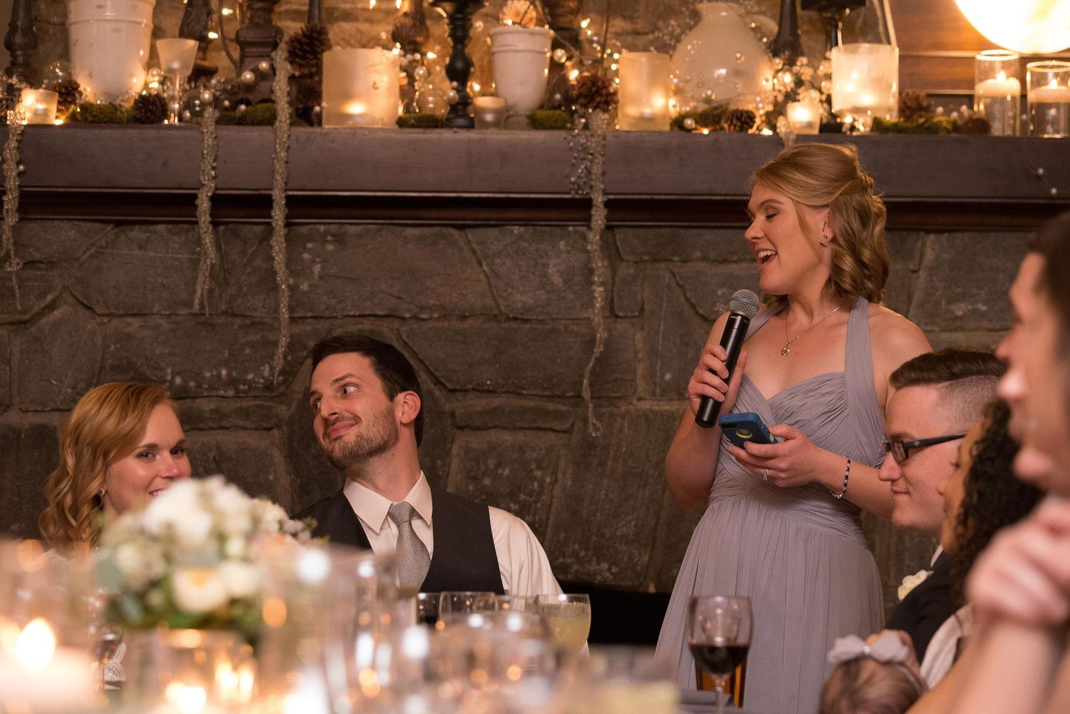Woman giving a speech at an elegant wedding reception dinner, holding a microphone near a stone fireplace.