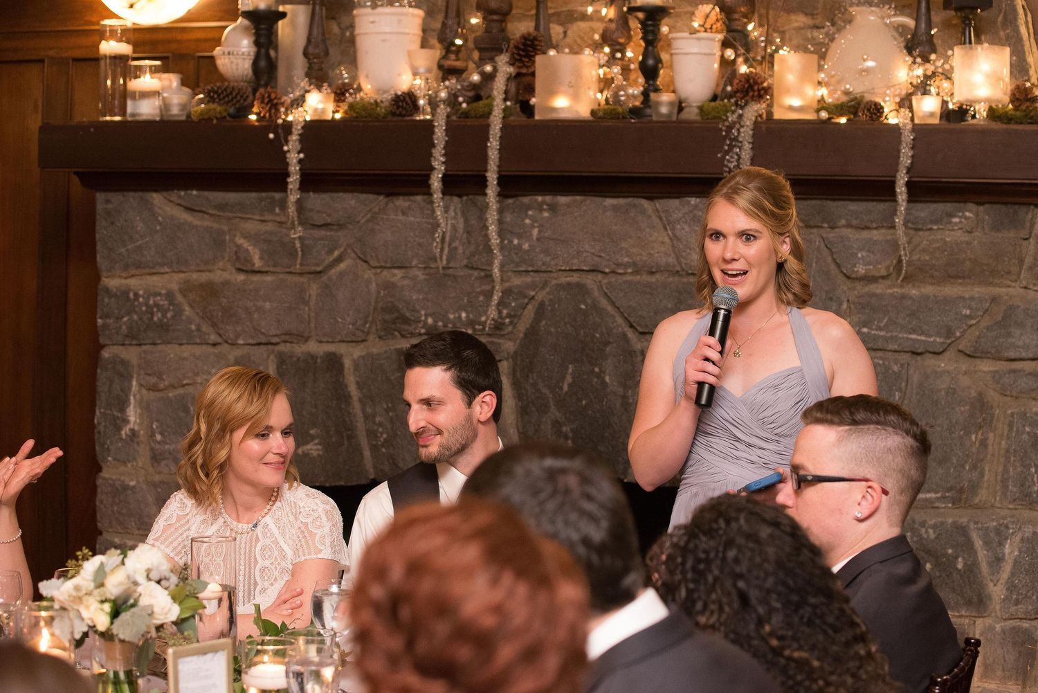 Woman giving a speech with microphone at a wedding reception near a stone fireplace with candles.