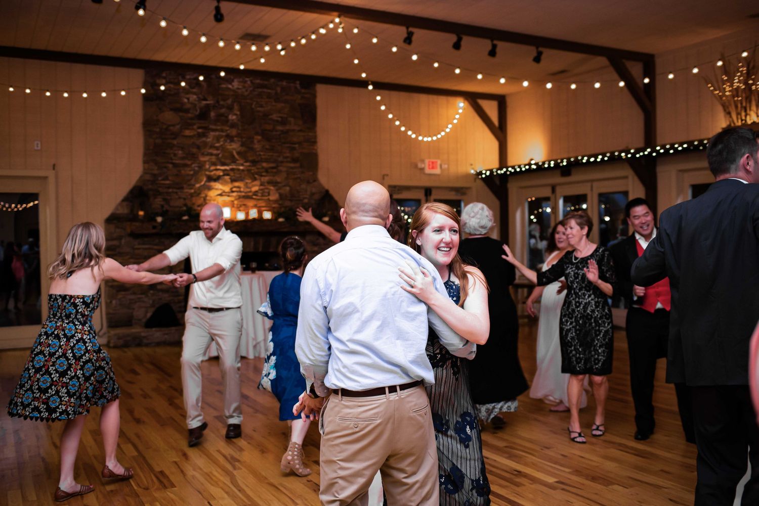 Guests dancing joyfully at a rustic wedding reception with string lights and stone fireplace backdrop.