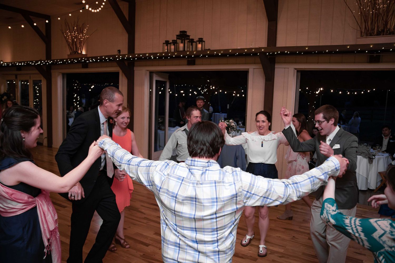 Wedding guests joyfully dancing in a circle at a barn reception venue decorated with string lights.