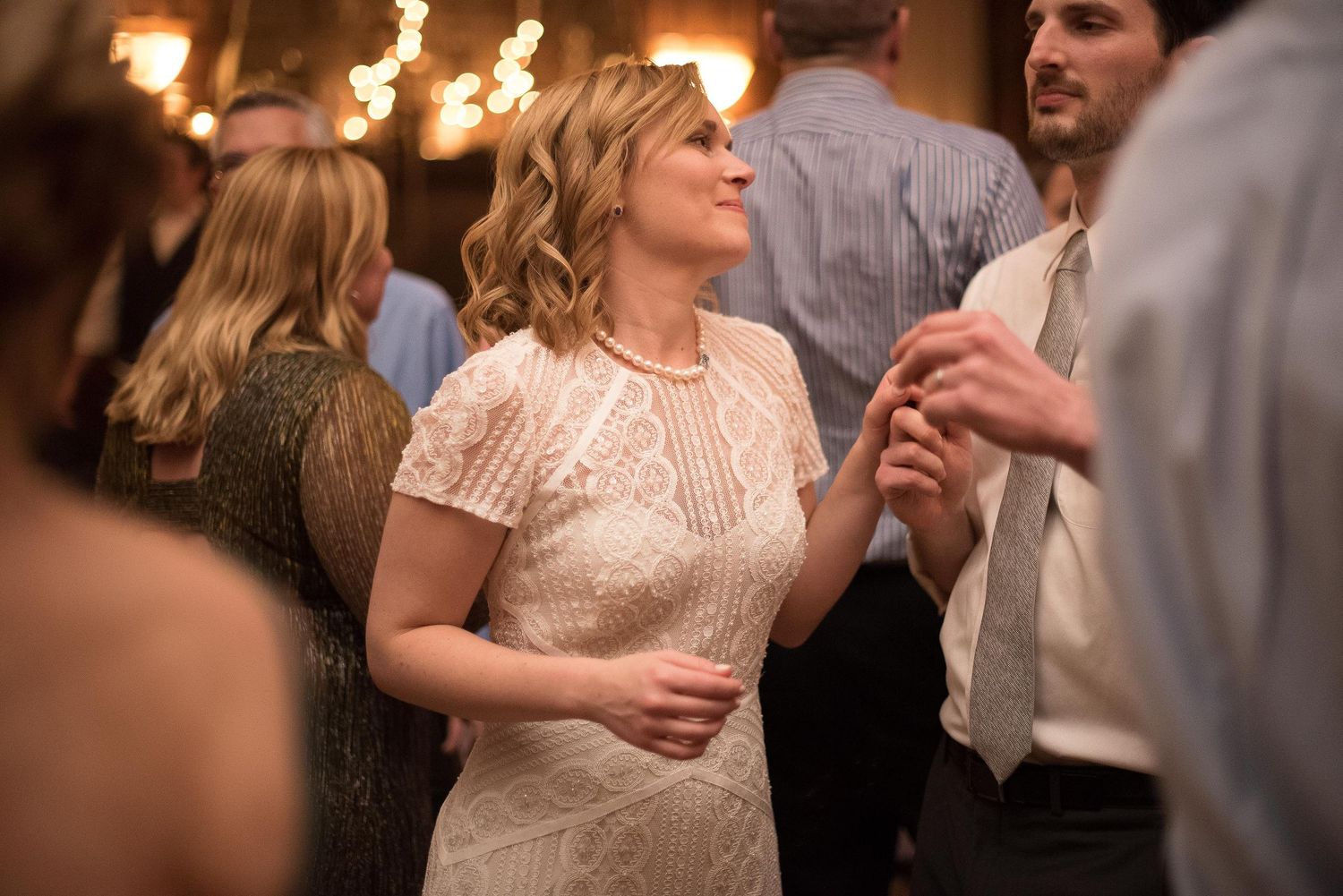 Woman in white lace dress dancing at an elegant wedding reception with warm string lights in background.