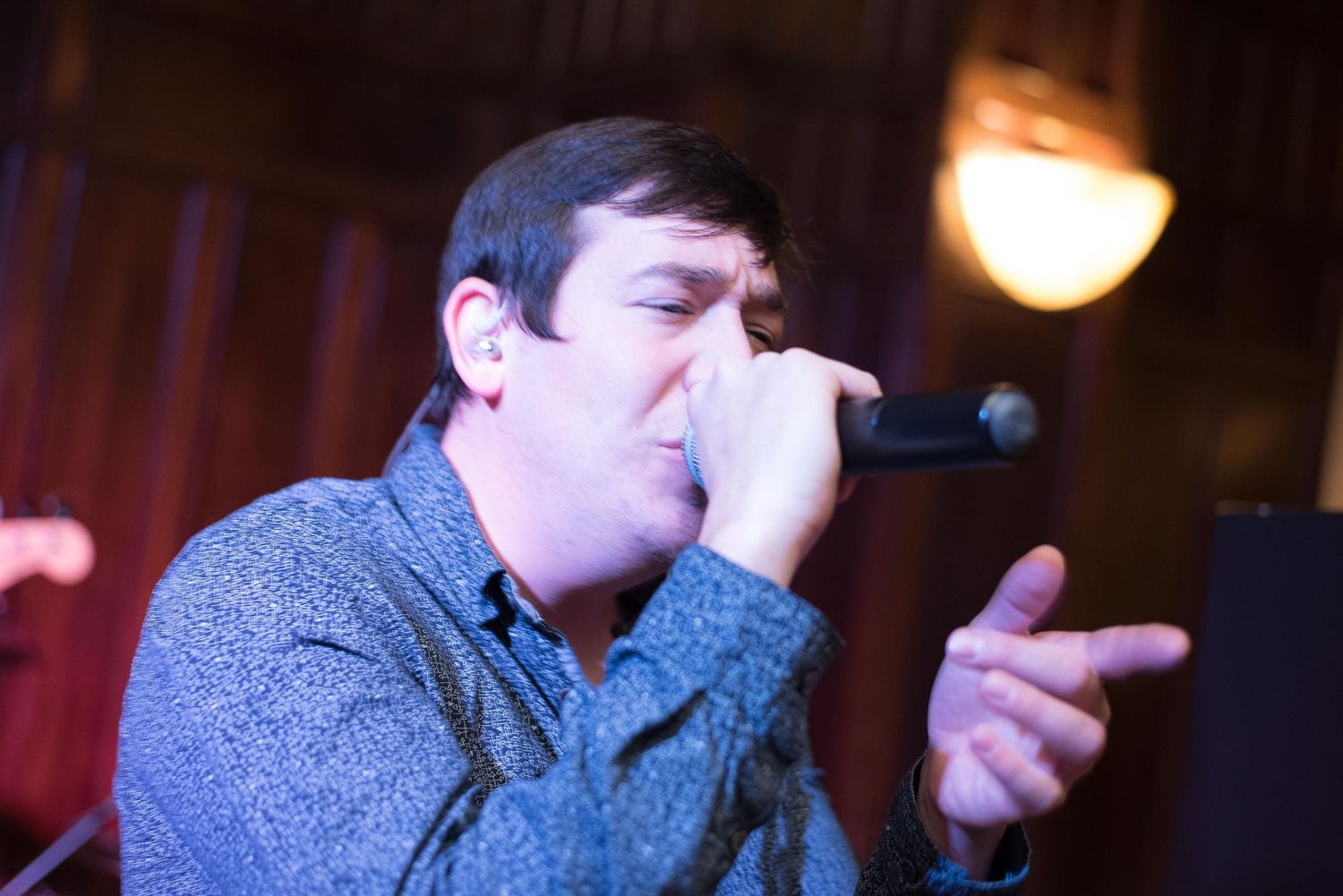 Man singing passionately into a microphone on stage, wearing a blue patterned shirt under warm venue lighting.