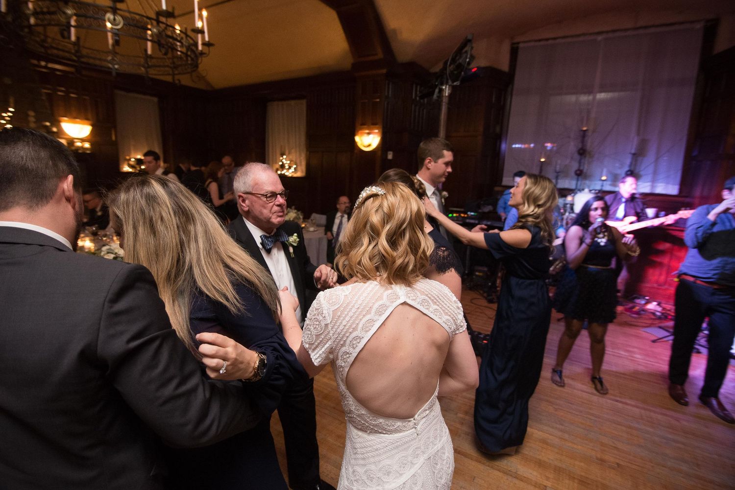 Bride and groom dancing with guests at elegant wedding reception in grand ballroom with chandelier lighting.