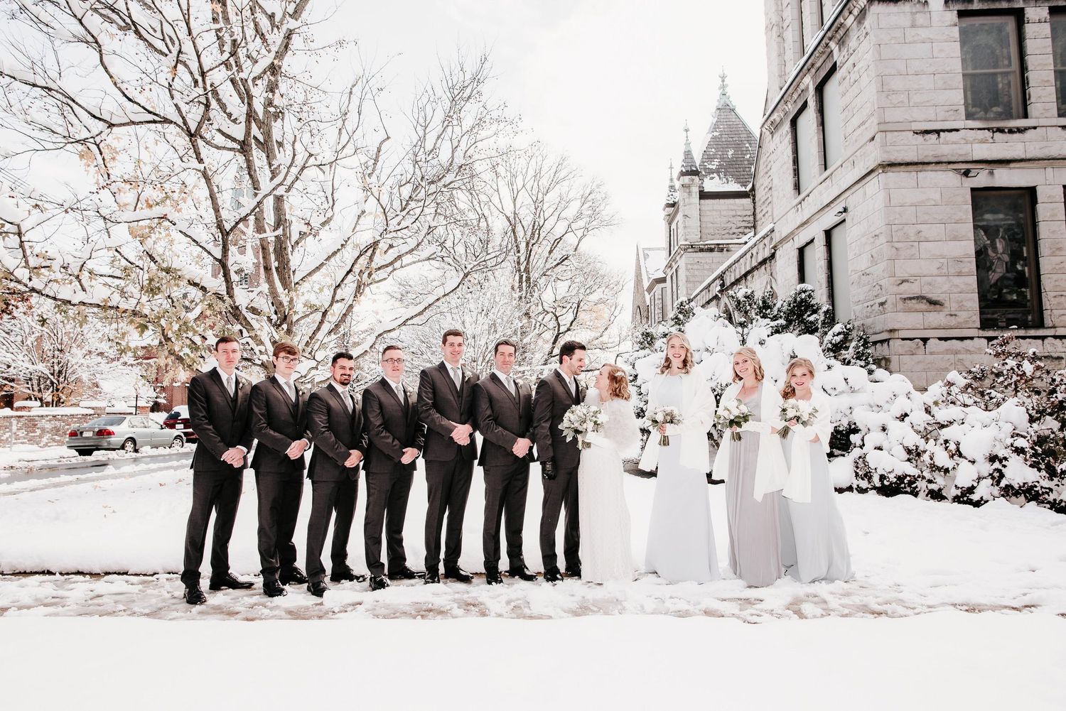 Wedding party posing in snow outside historic stone building, groomsmen in black suits and bridesmaids in silver gowns.
