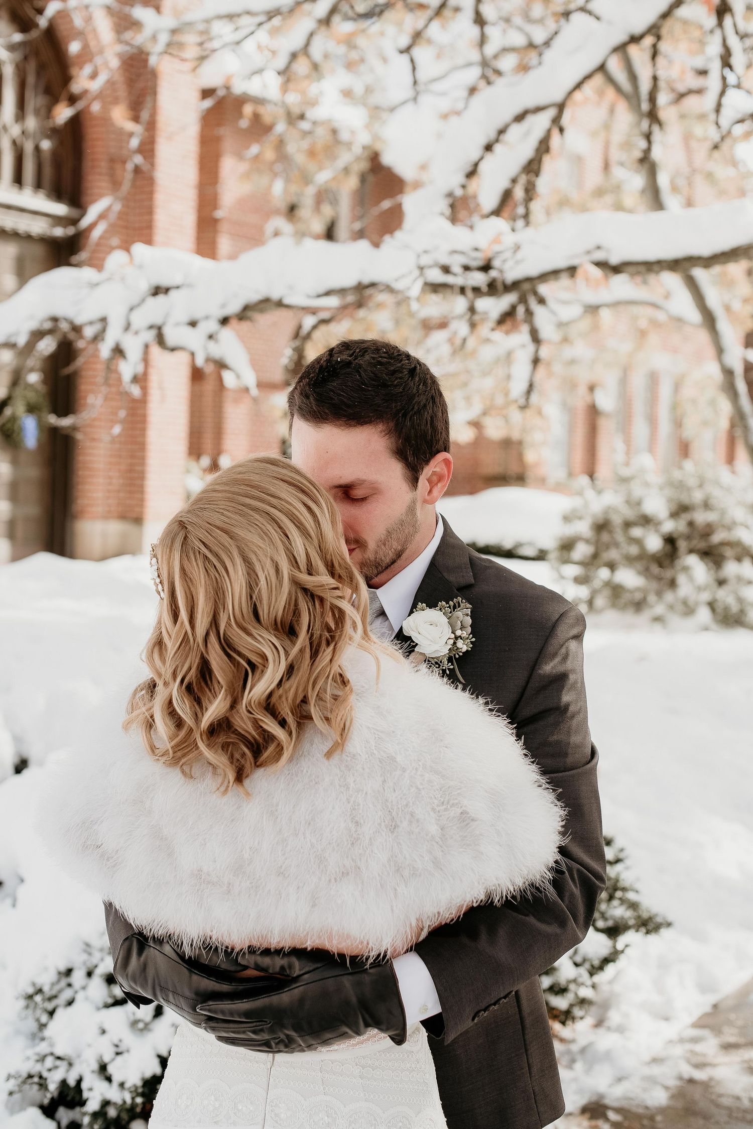 Bride and groom sharing a kiss in snowy winter setting, bride wearing white fur wrap and groom in dark suit with boutonniere.