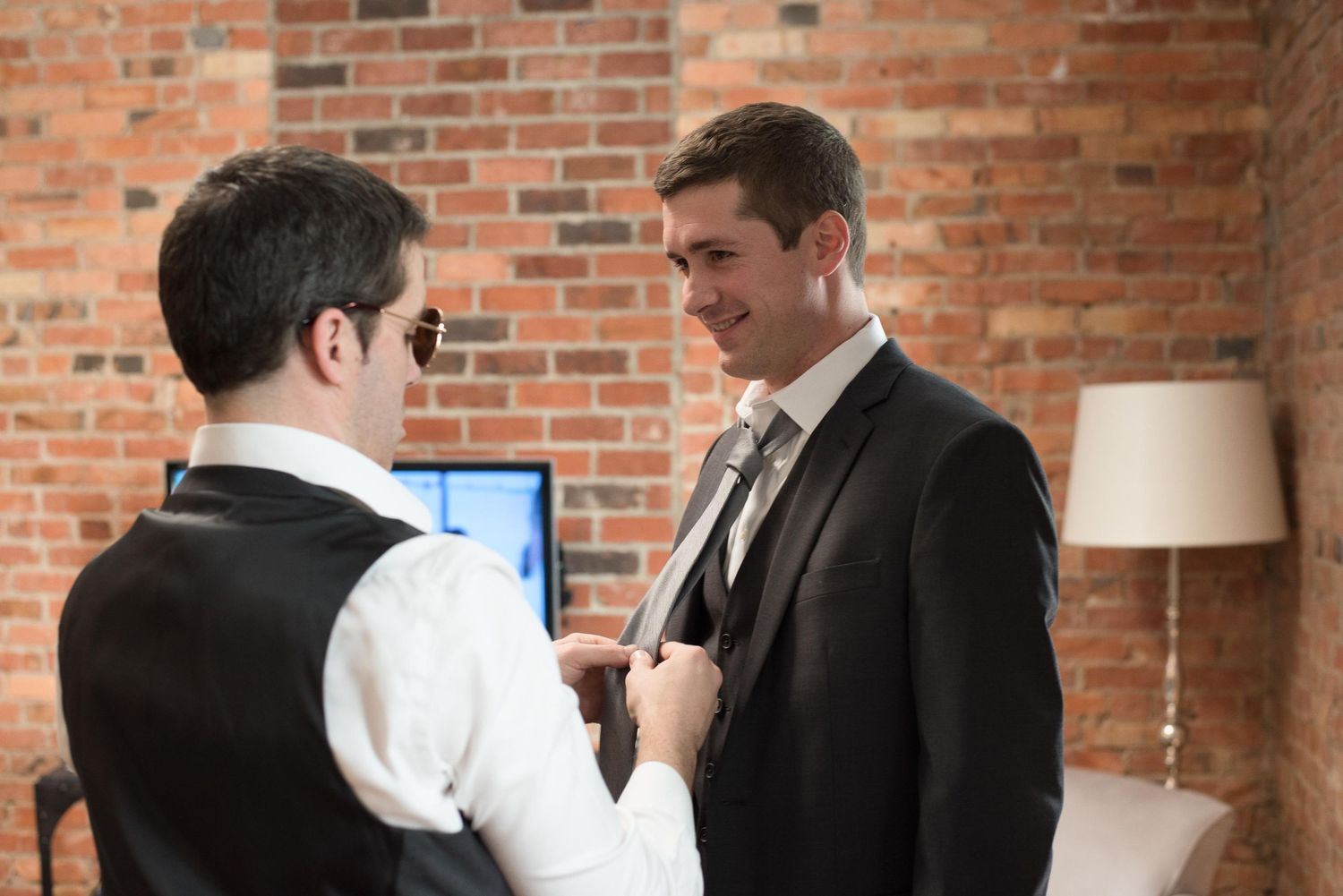 Two men in formal suits share a moment as one adjusts the other's tie in a brick-walled room.
