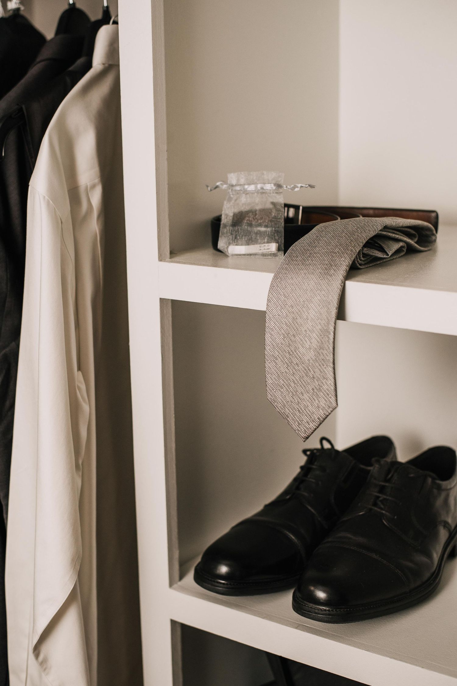 White closet shelves displaying black leather oxford shoes, a gray tie, wallet, and cologne beside hanging suits.