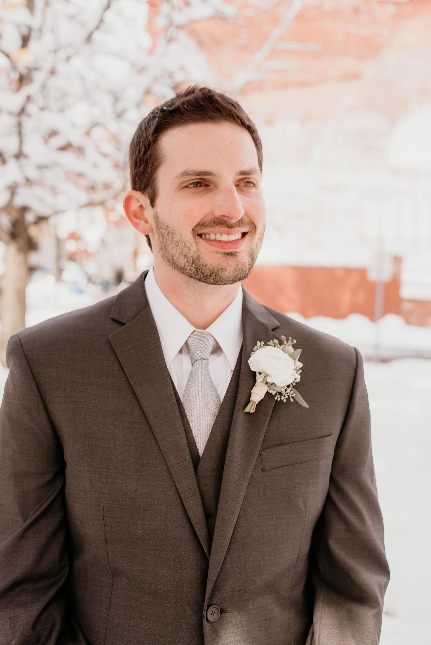 Smiling groom in gray suit with white boutonniere poses outdoors in snowy winter wedding setting.