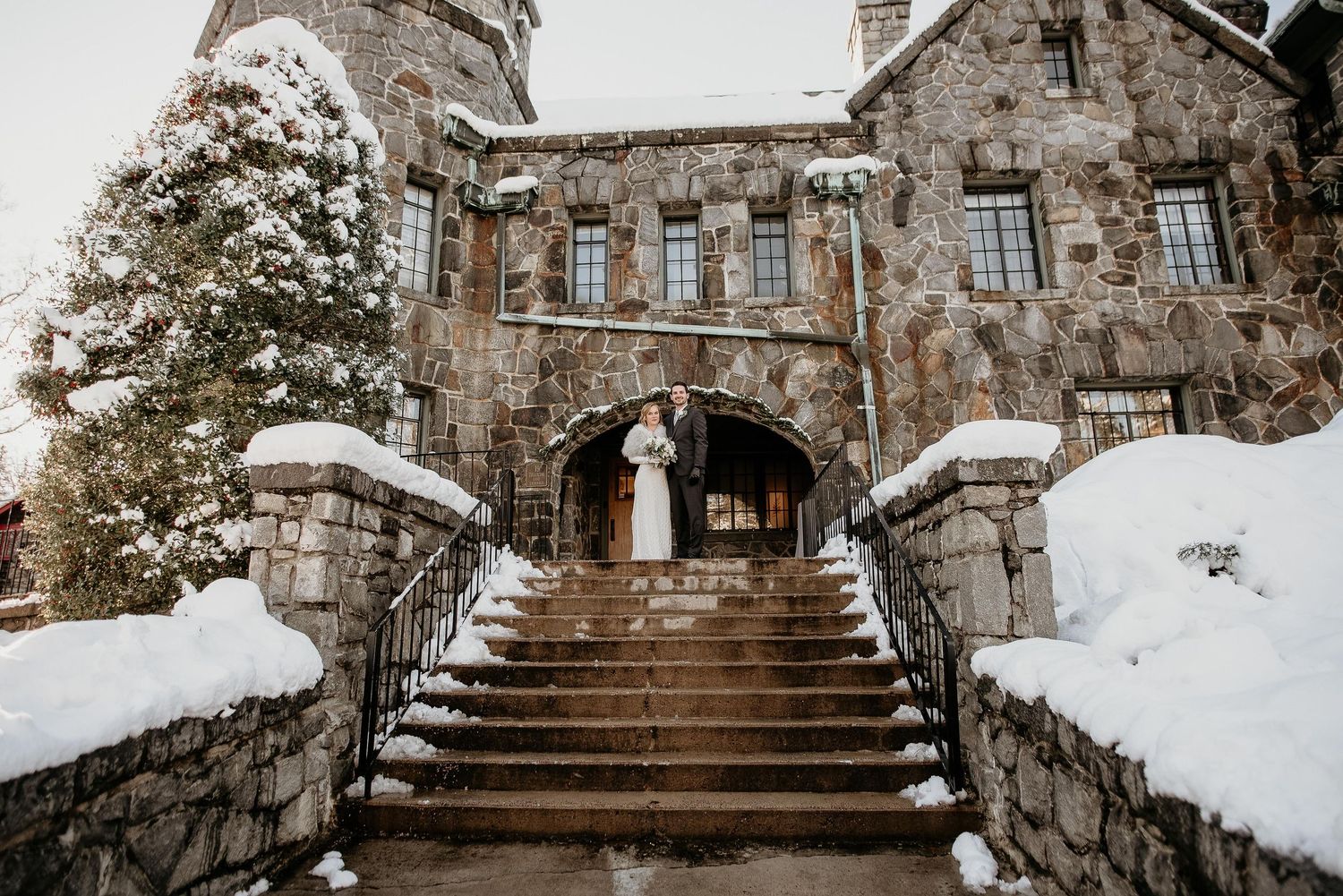 Couple embracing on snow-covered stone castle steps in winter wedding attire.