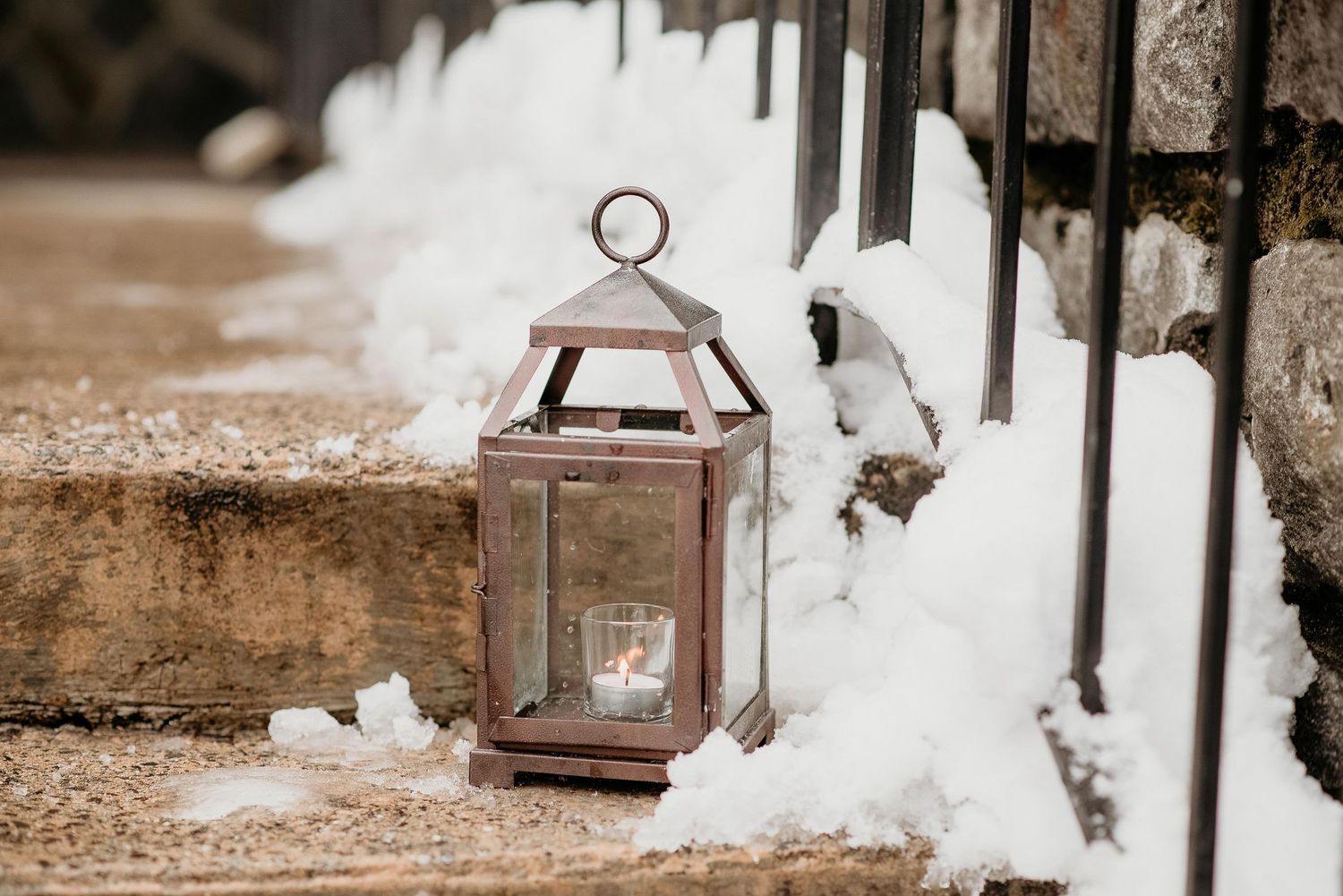 A rustic metal lantern with a glowing candle sits on snow-dusted stone steps beside a black iron fence in winter.