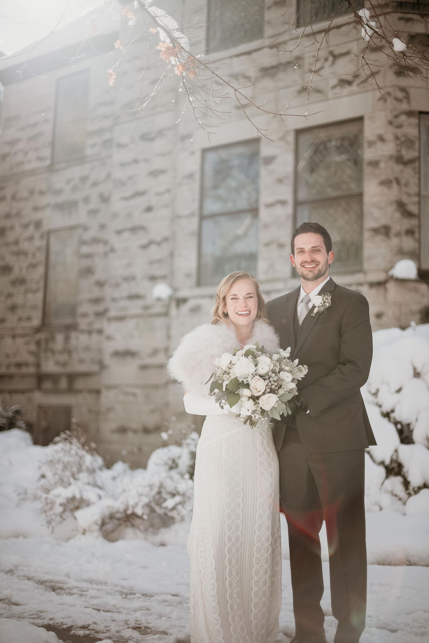 Bride and groom smiling together in winter snow, bride holding white bouquet wearing fur wrap outside stone building.