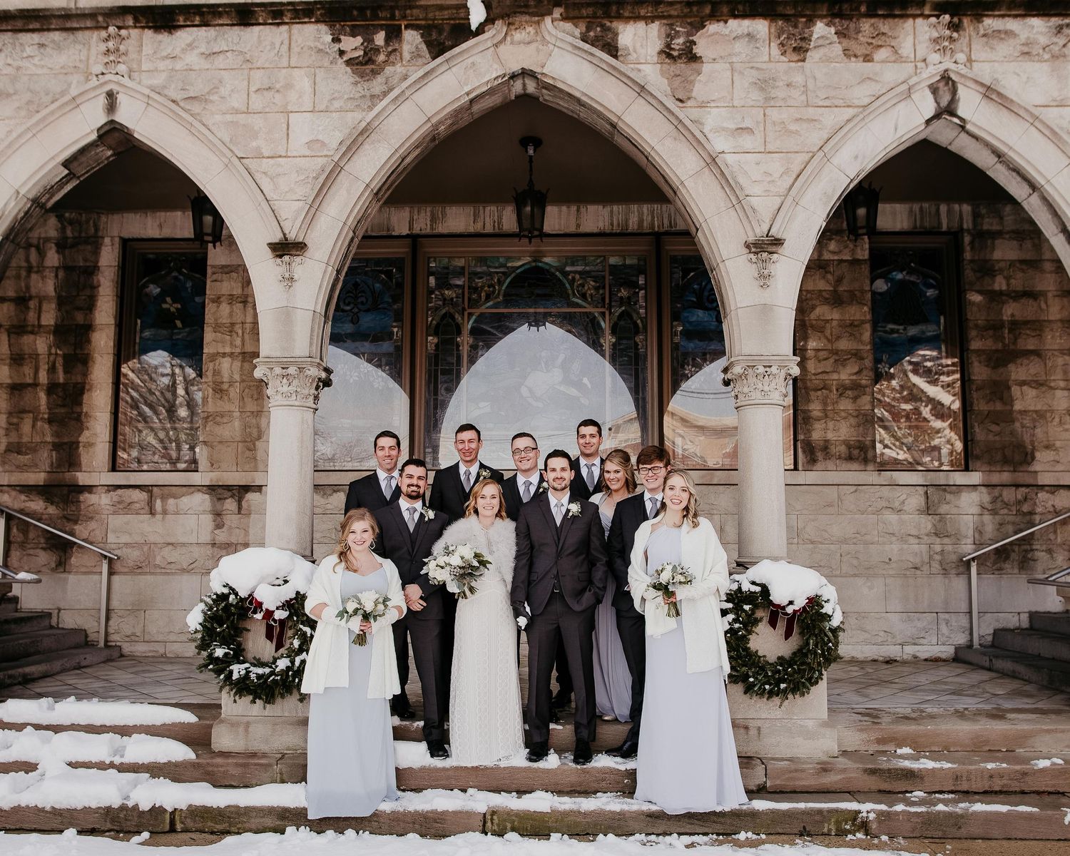 Wedding party posing on snowy church steps in white and gray attire with floral wreaths.