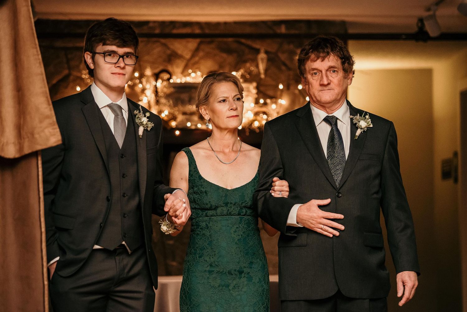 Family of three posing elegantly at a formal event, woman in green dress flanked by two men in dark suits.