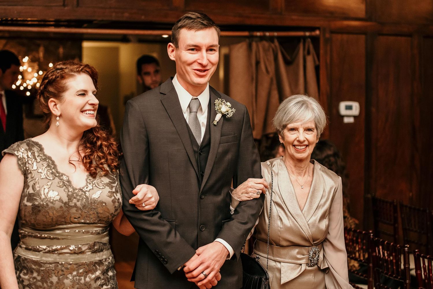 Groom in gray suit escorted by two women in elegant dresses at a wedding reception venue.