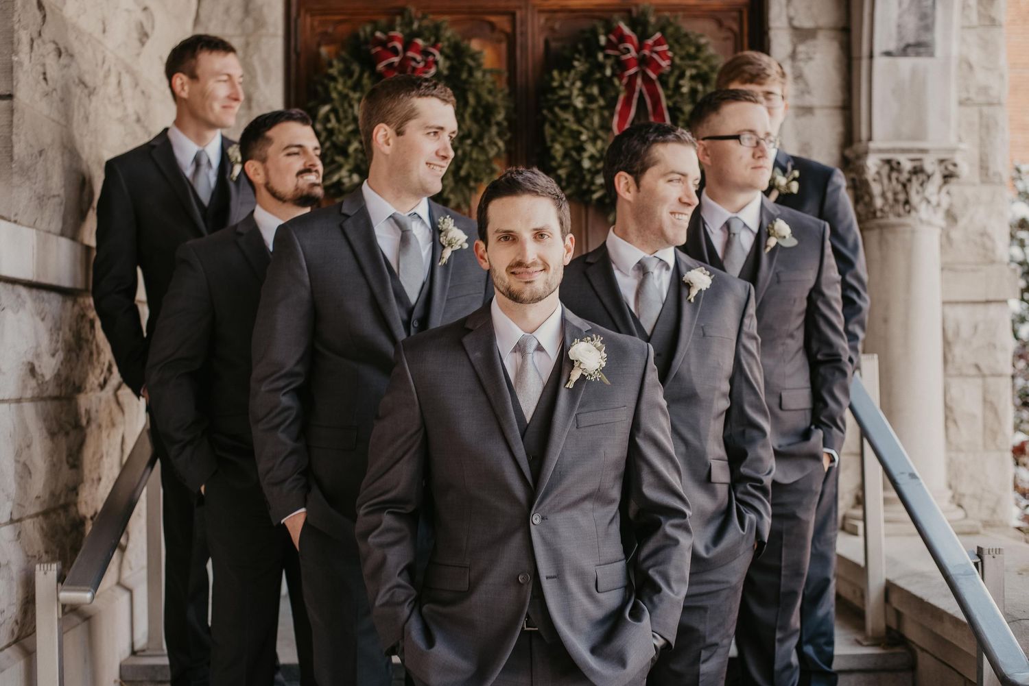 Groom and groomsmen in charcoal suits with boutonnieres posing on stone steps with holiday wreaths backdrop.