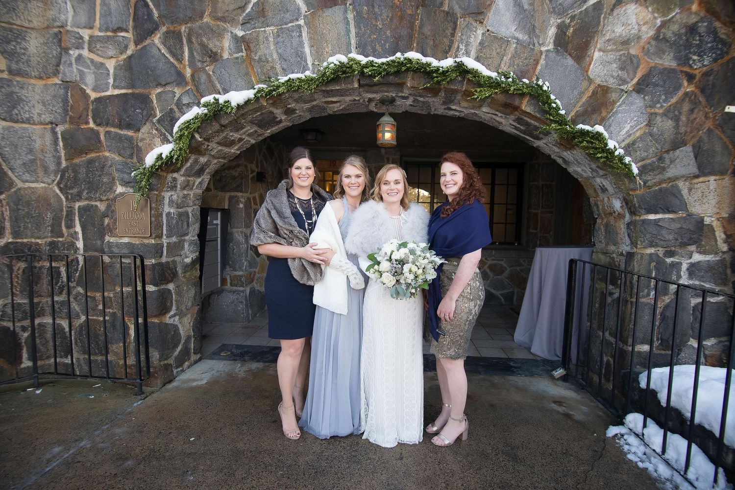 Bride holding bouquet poses with three bridesmaids in front of stone archway at winter wedding venue.